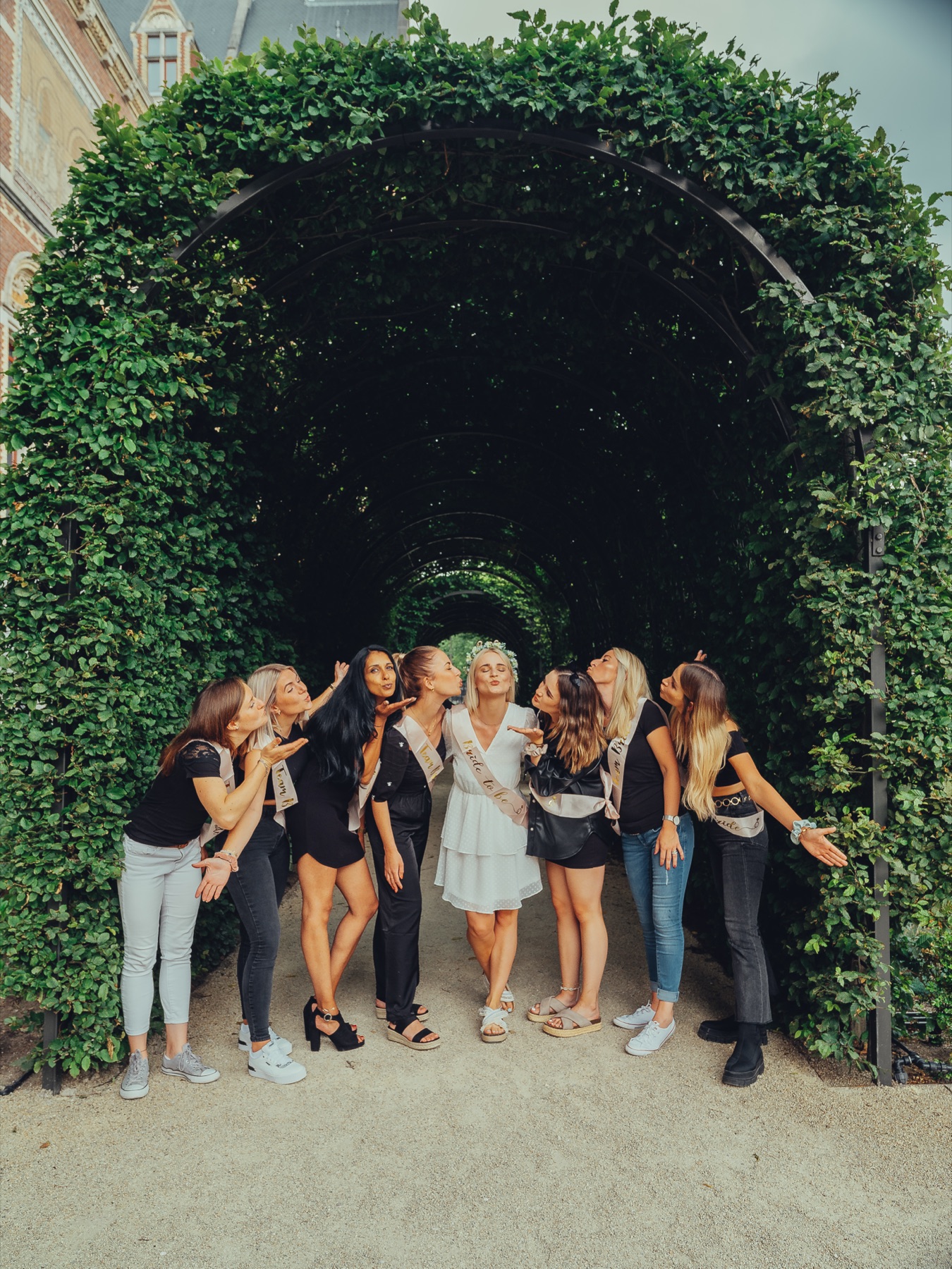 Bachelorette friends blowing kisses in a long arch of green hedges beside the red brick of the Rijksmuseum