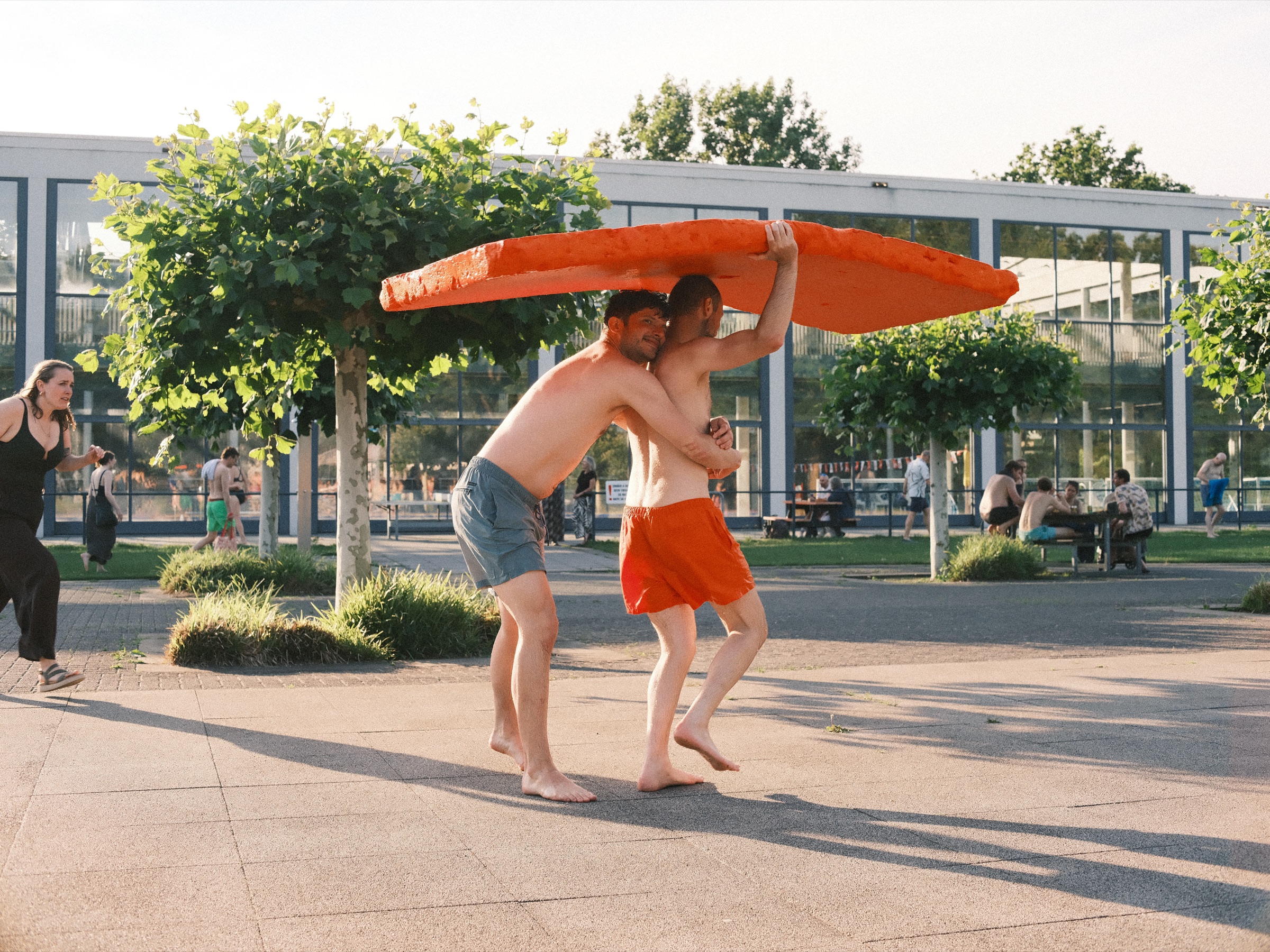 Two friends in swim shorts carrying an orange mat over their heads past a glass pool building, long shadows on the tiles