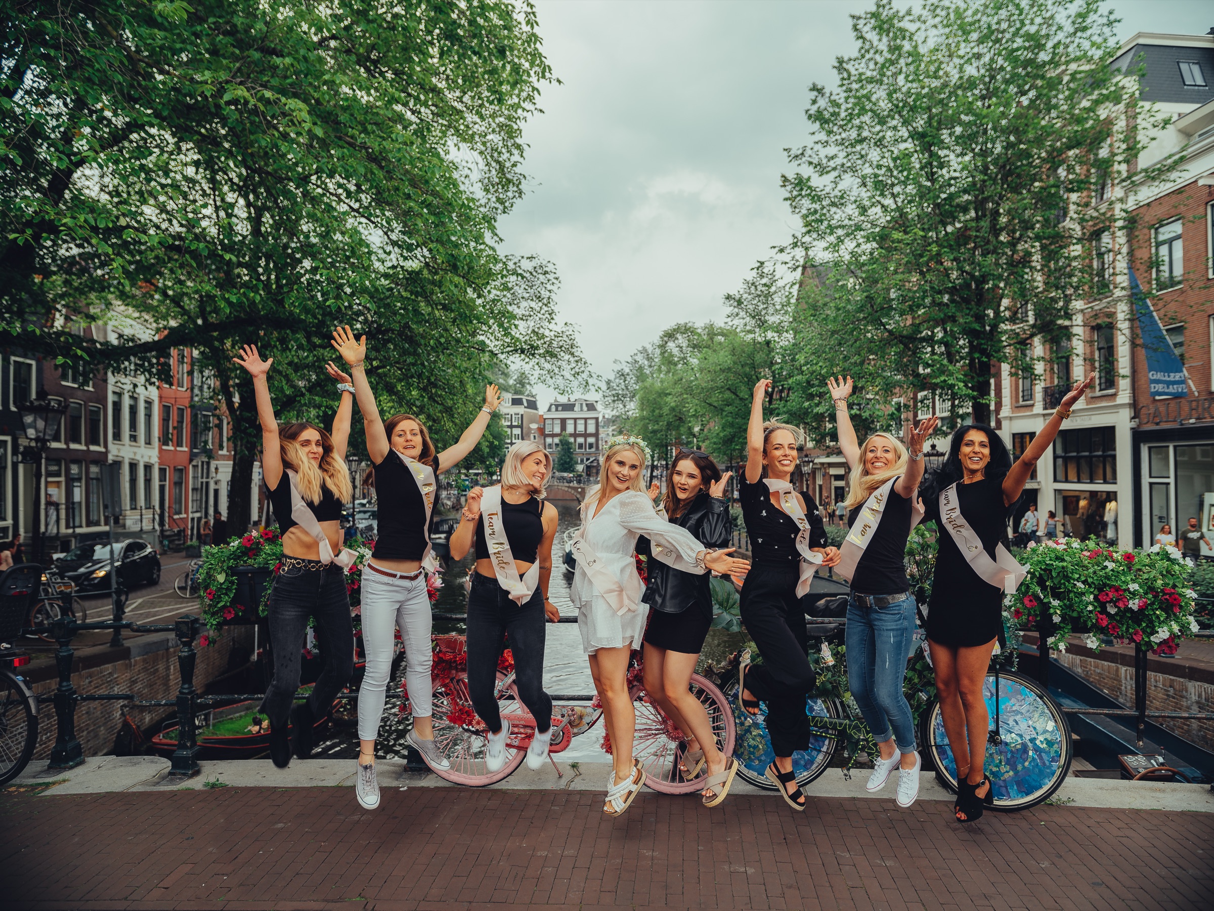 Friends jumping on a canal bridge in front of flower-covered bikes, the bride in white in the middle of the group
