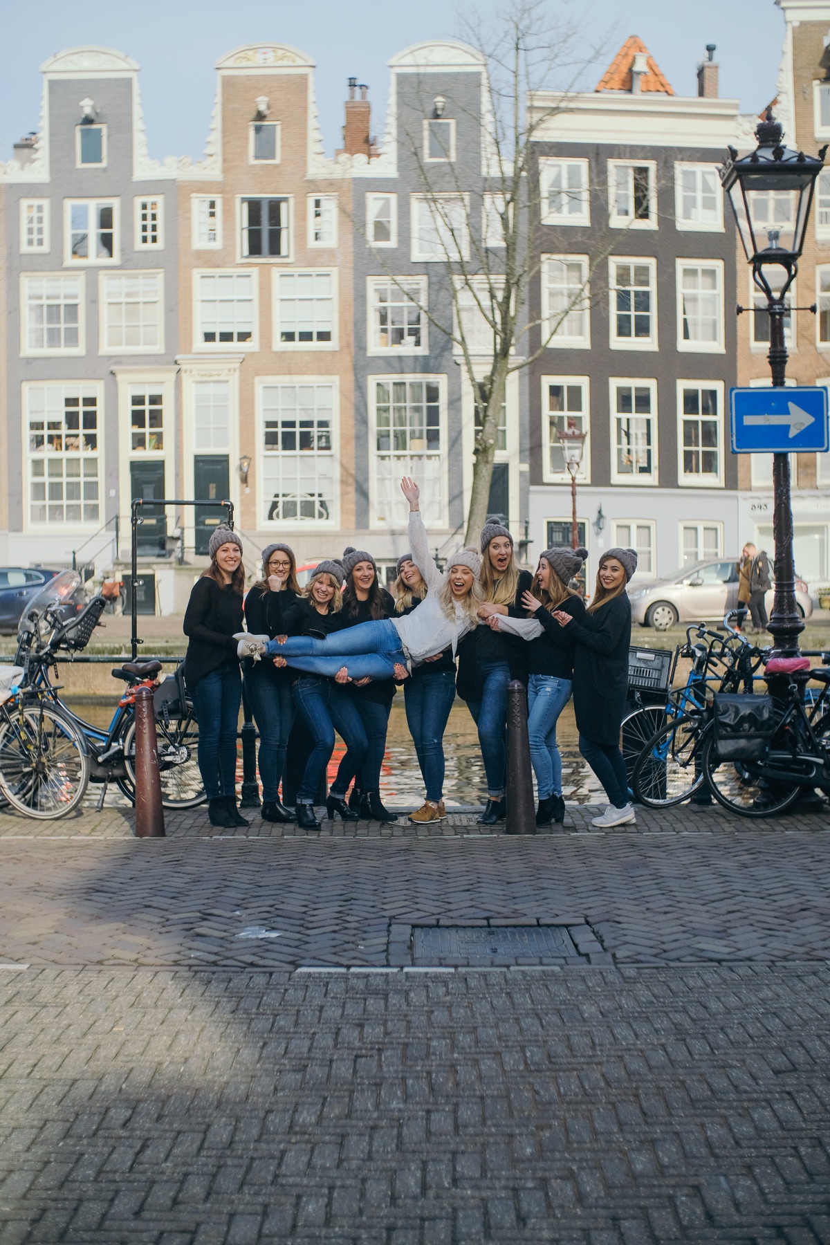 Hen party friends lifting the bride sideways beside a canal, gabled houses and an old lamp post with a blue arrow sign behind