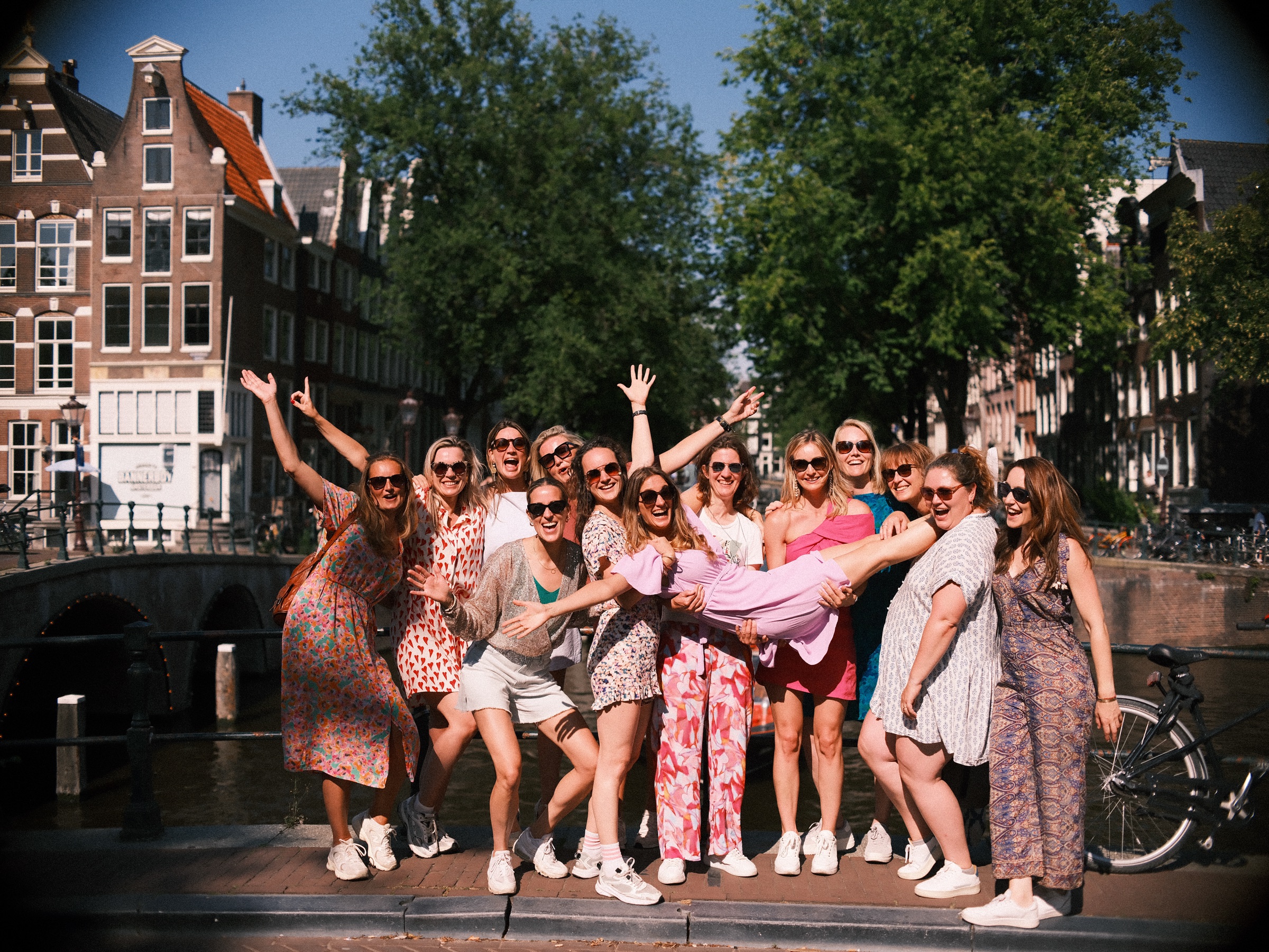 Twelve friends in summer dresses cheering on a canal bridge as they hold the bride-to-be across their arms at her bachelorette