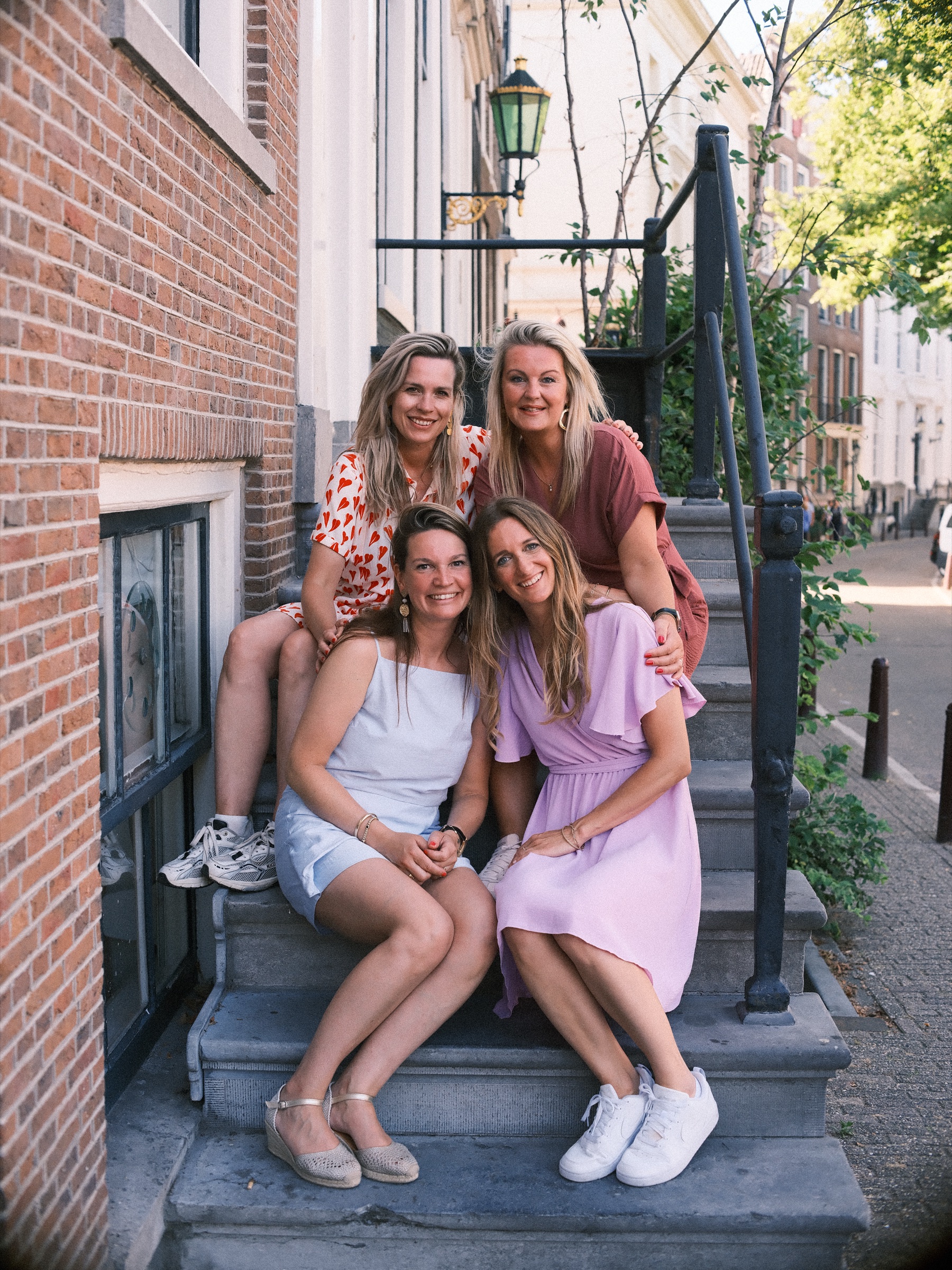 Four women sitting together on the stone steps of a canal house stoop, arms around each other and laughing