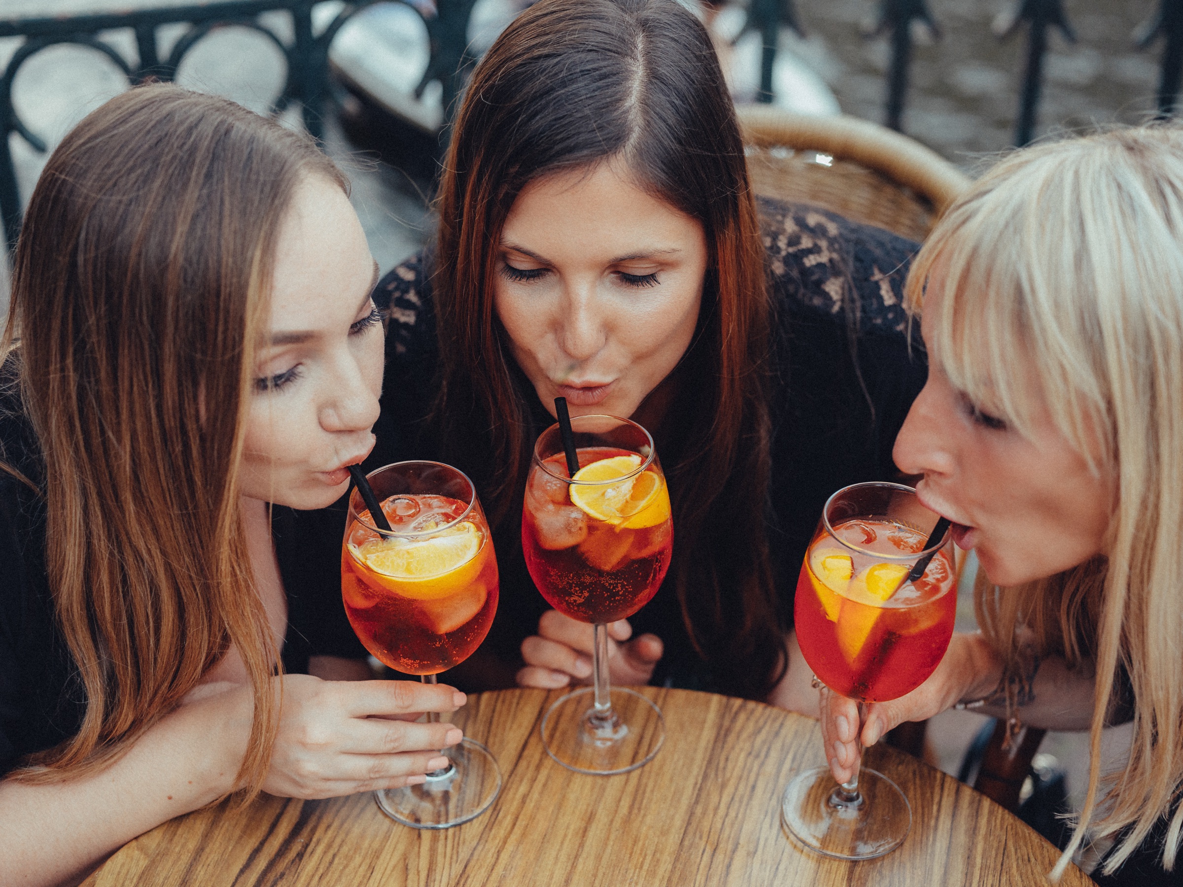 Three friends sipping spritz through straws at a round café table by the canal, iron railings and boats behind them
