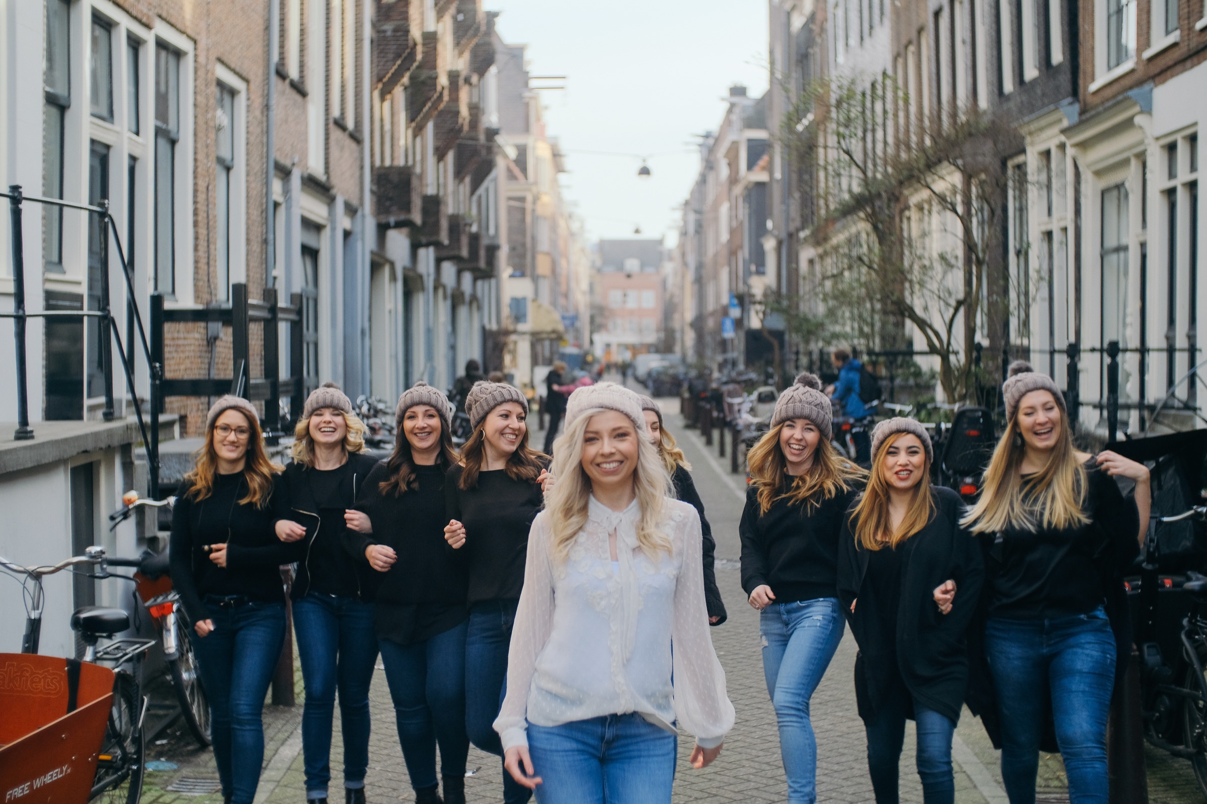 Friends in black tops and gray beanies walking arm in arm down a bike-lined street, the bride in white a step ahead