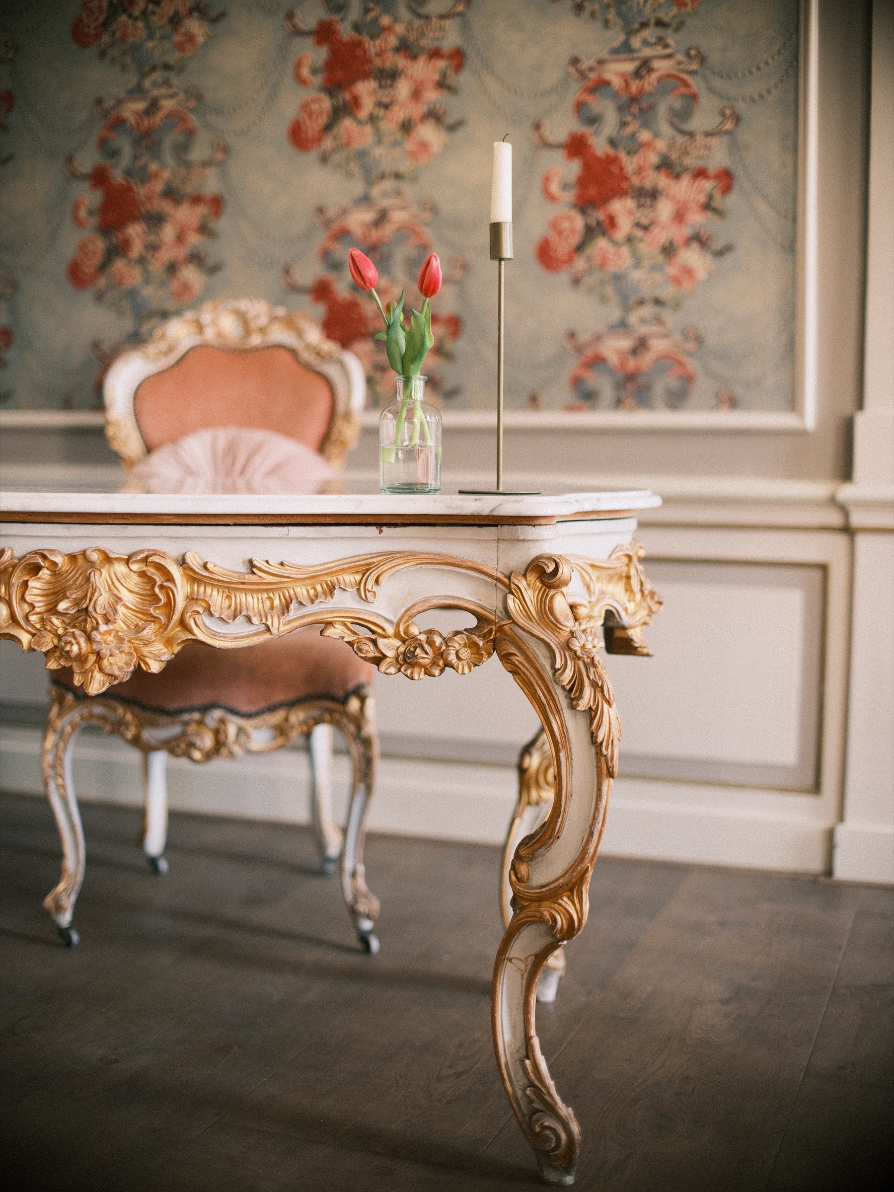 Vergulde tafel met een wit marmeren blad, een roze fluwelen stoel en rode tulpen in een glazen vaasje voor bloemenbehang