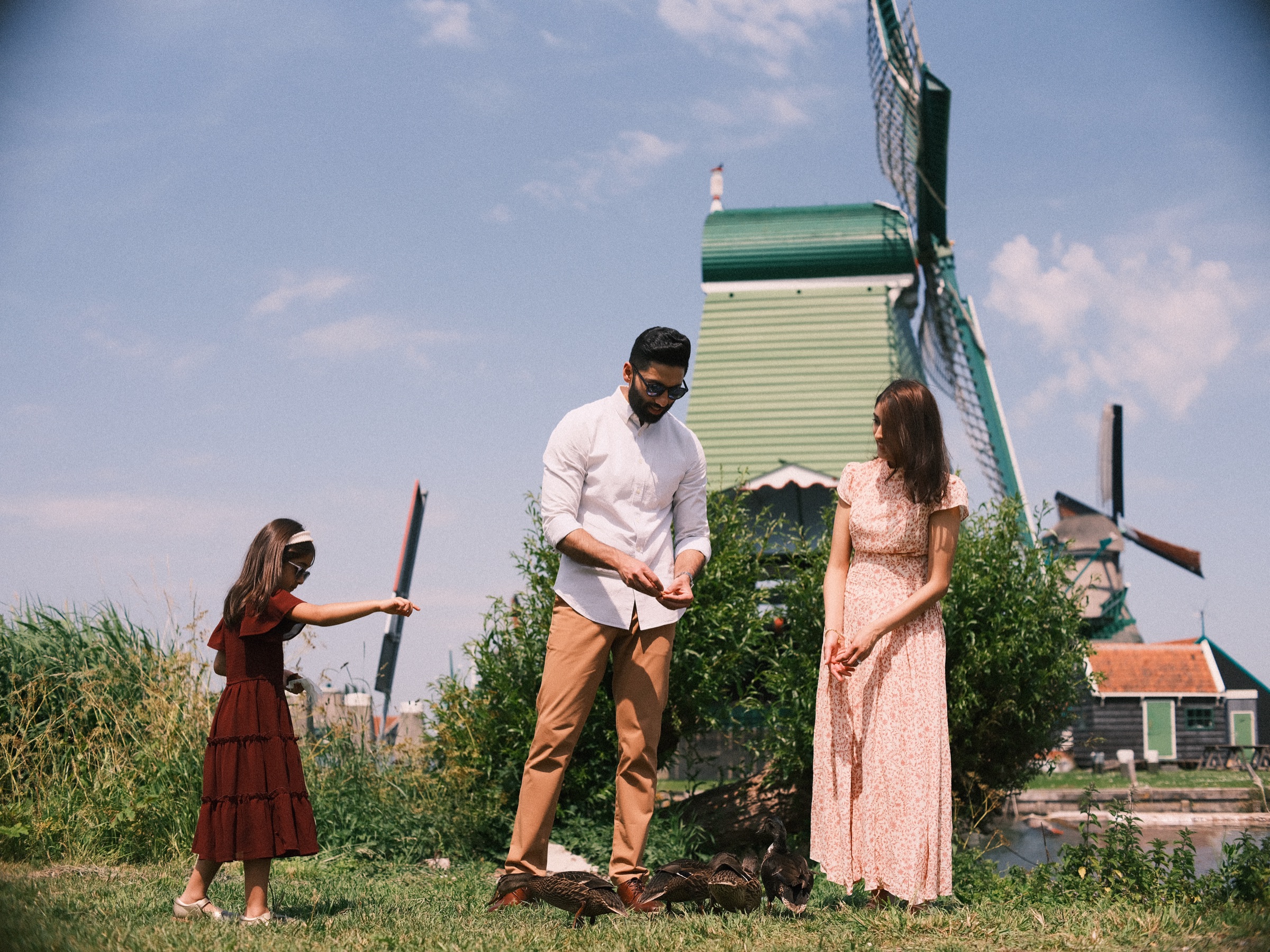 Girl in a dark red dress reaching out to the ducks while her father feeds them by hand and her mother looks on beside the mill