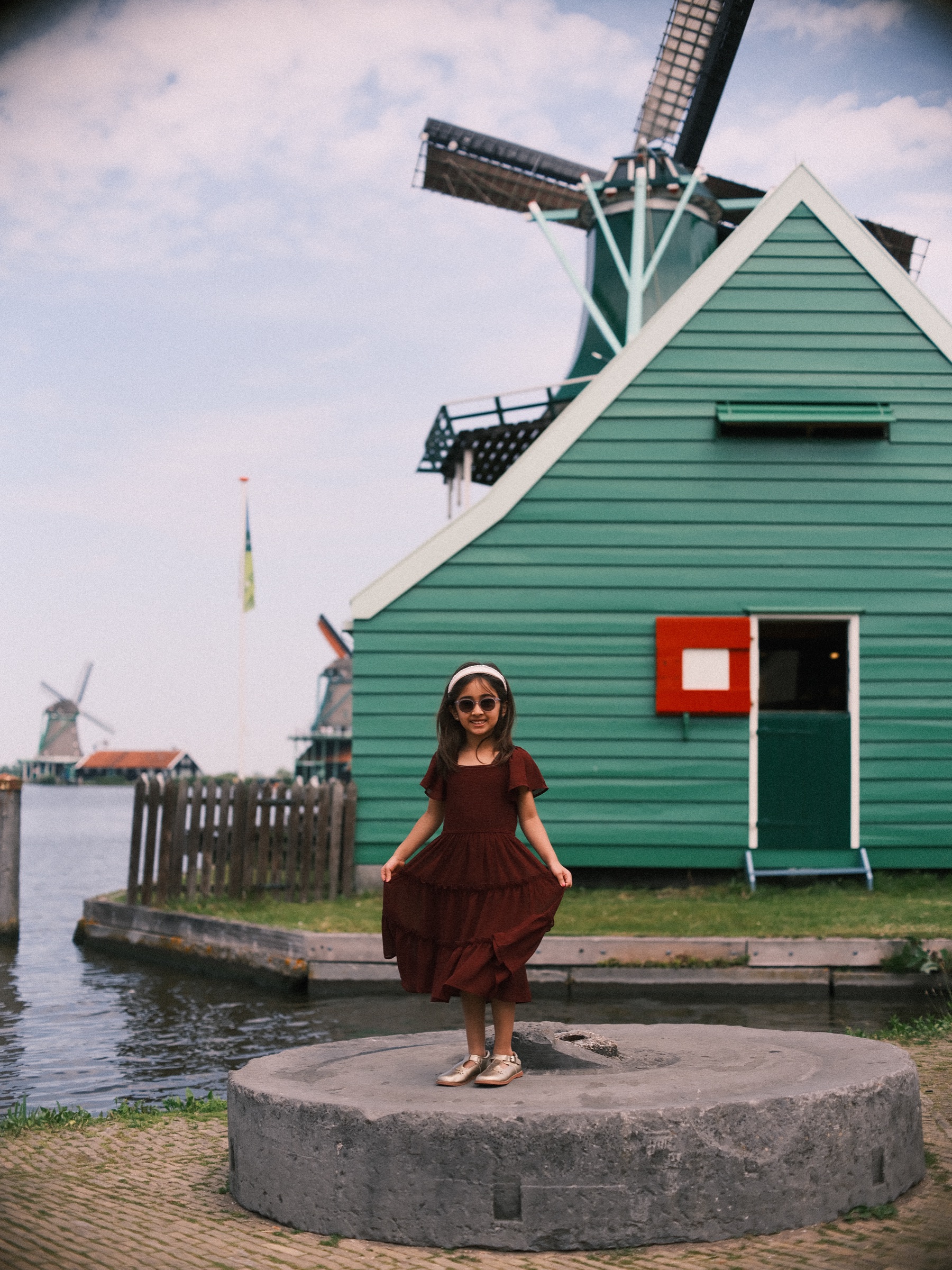 Little girl in a dark red dress and white sunglasses holding out her skirt on an old millstone in front of a green wooden house
