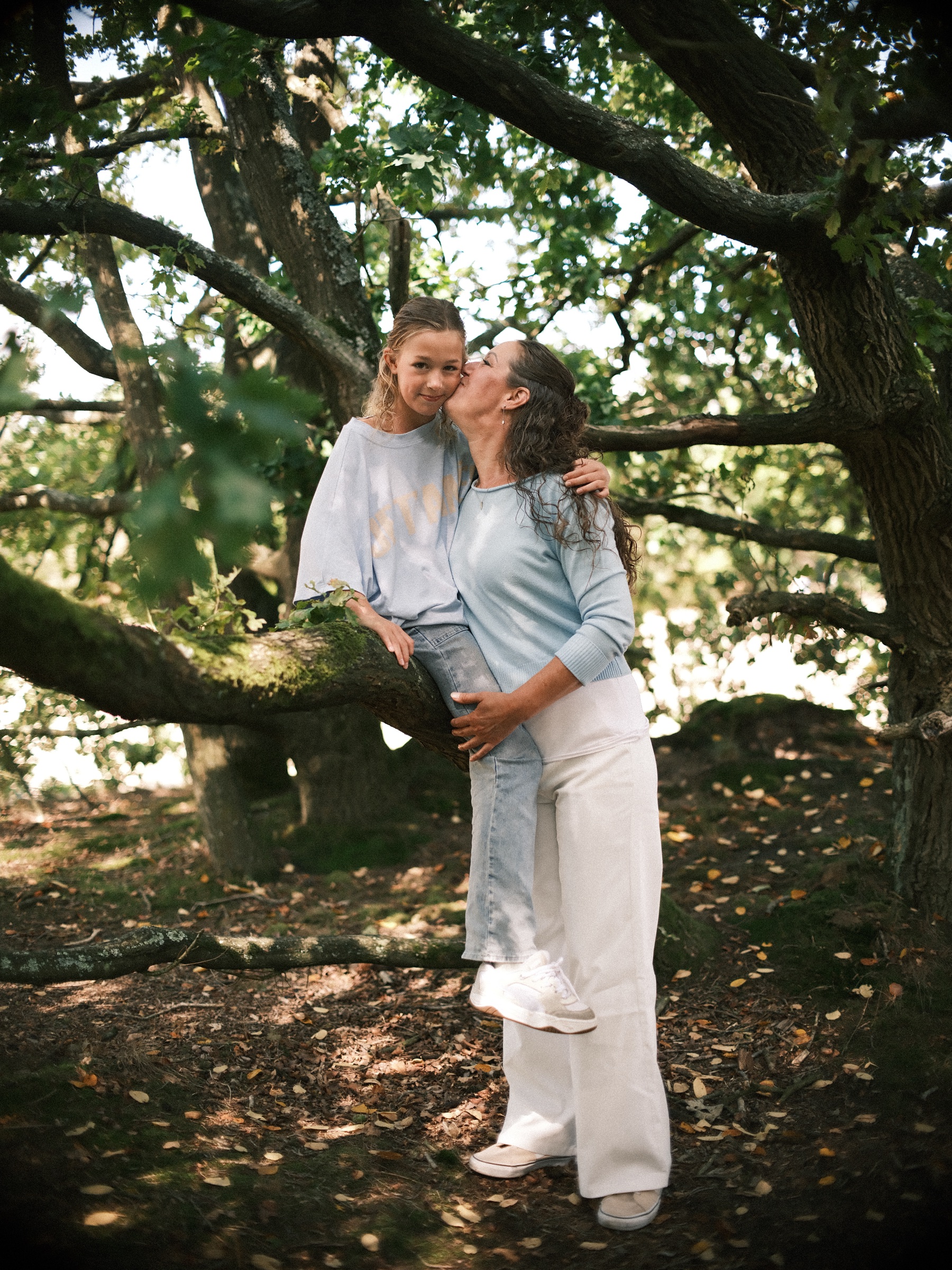 Girl sitting on the low branch of an old oak while her mother kisses her cheek, sunlight falling through the green leaves