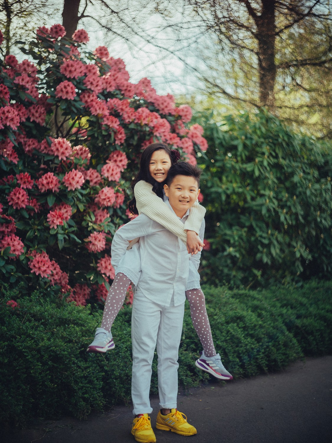 Meisje zit lachend op de rug van haar broer langs roze rododendrons, allebei in het wit gekleed