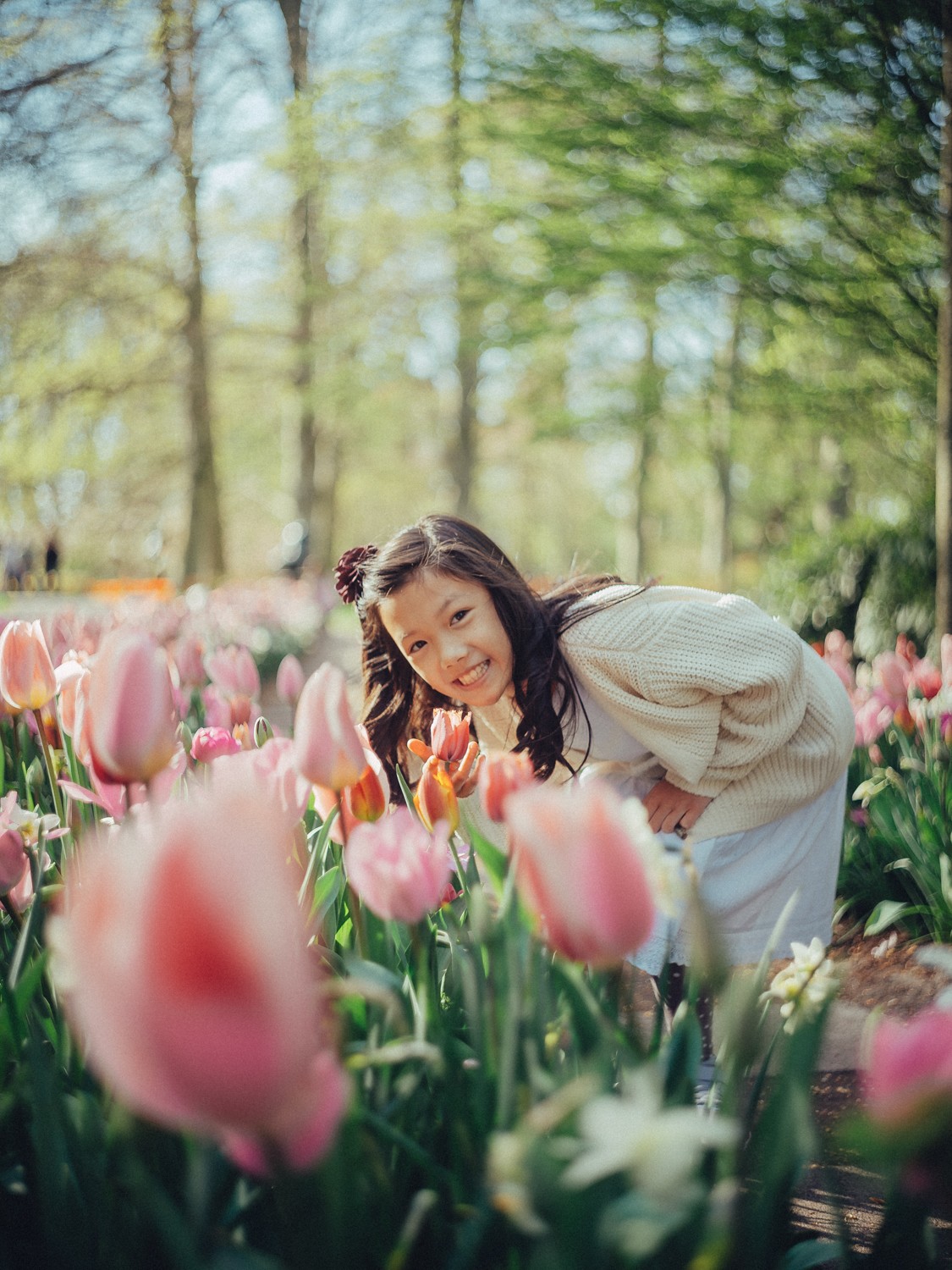Meisje in een crèmekleurig vest buigt zich tussen roze tulpen en grijnst naar de camera, de jongste generatie in Keukenhof