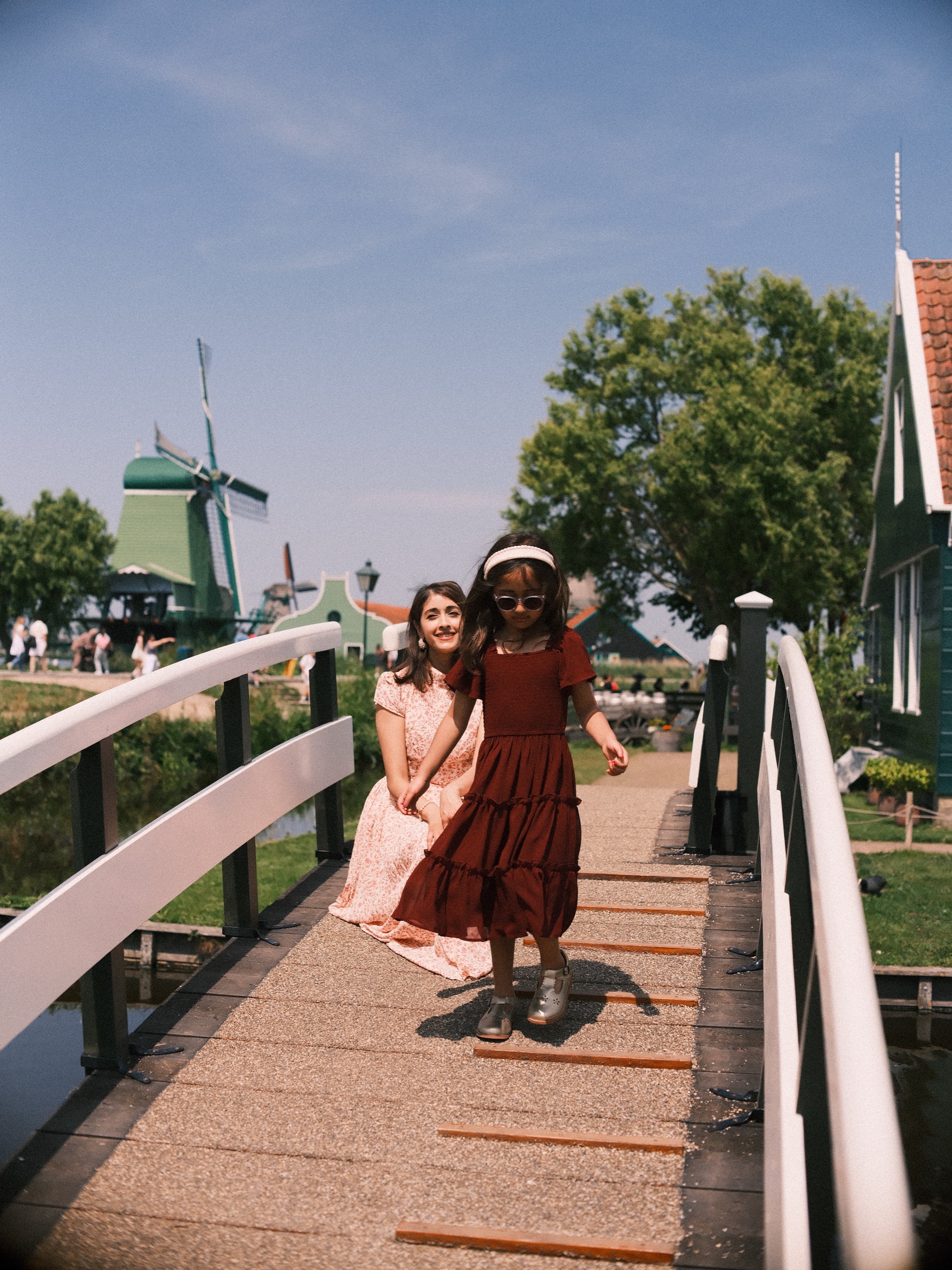 Little girl skipping across a white bridge in front of her smiling mother, green houses, trees and a windmill in the distance