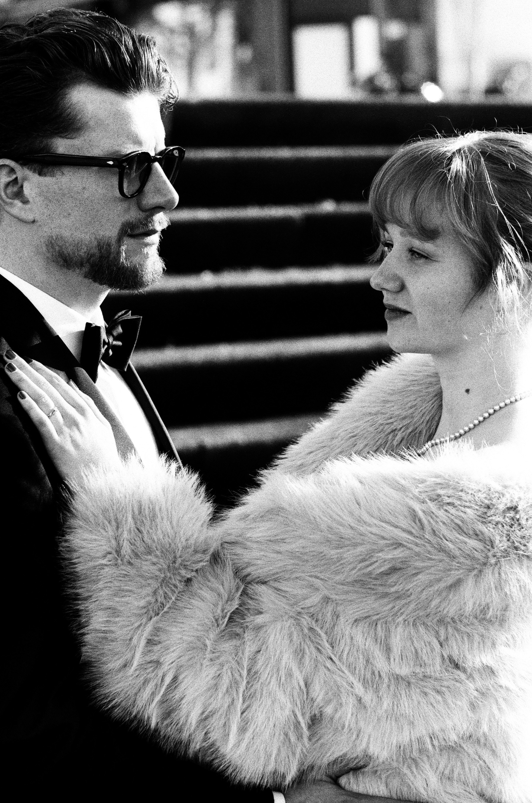Close view of the two of them face to face, his glasses and bow tie, her fur coat and pearls, stone steps behind them in black and white