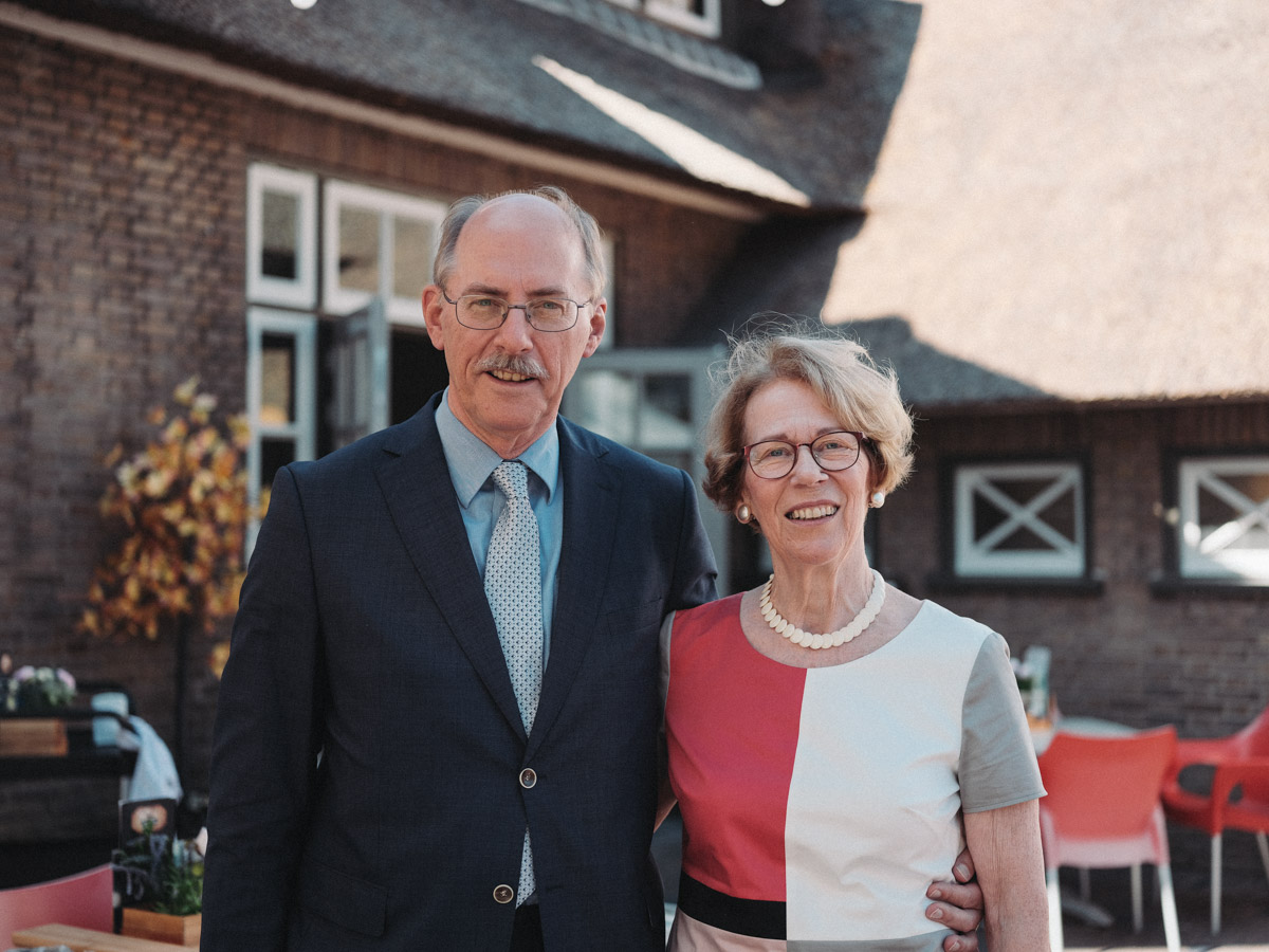 Couple smiling arm in arm on the terrace of Het Witte Paard on their golden anniversary in Rotterdam, she in pearls and a color-block dress
