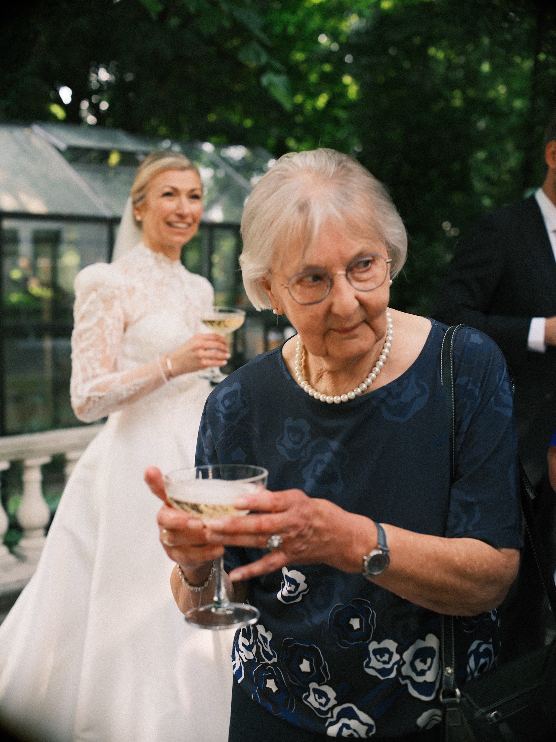 Oudere vrouw met parels houdt een glas champagne met twee handen vast, terwijl de lachende bruid met haar eigen glas achter haar staat