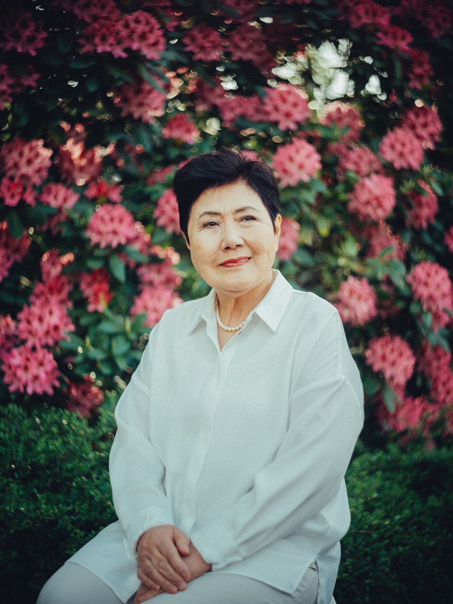 Oma in een witte blouse met parelketting zit voor een muur van roze rododendrons, de oudste generatie in Keukenhof