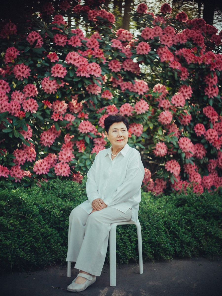 Oma zit op een wit krukje in een witte blouse en broek, omlijst door een hoge muur van roze rododendronbloemen