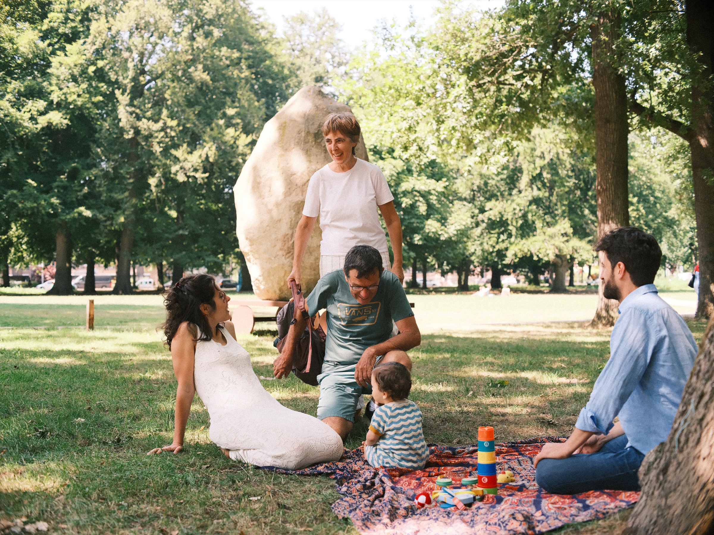 Opa en oma komen aan bij de picknick, opa hurkt bij de baby terwijl oma glimlachend voor een grote kei staat