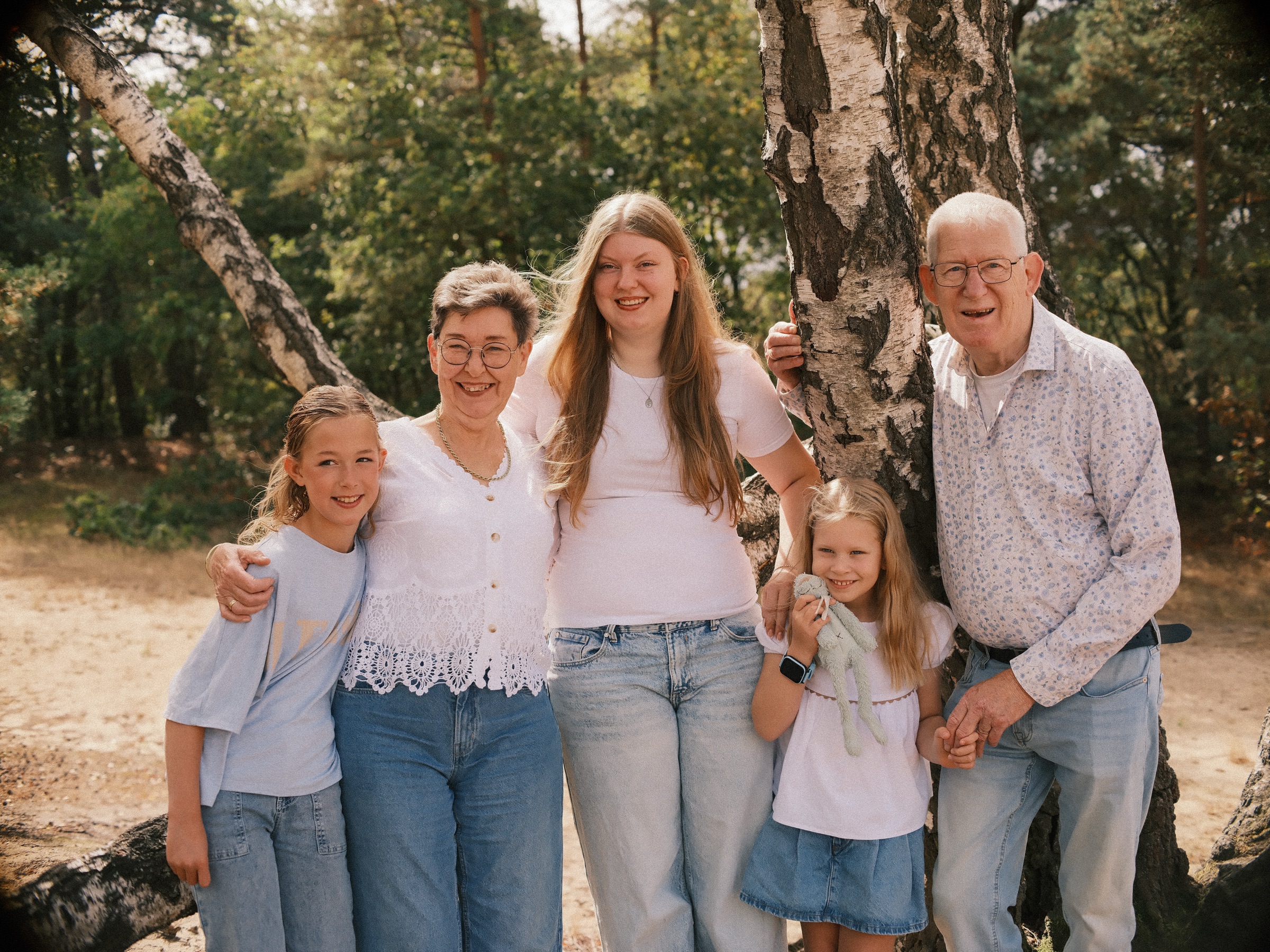 Grandmother and grandfather with three granddaughters by a birch tree, the youngest holding a soft toy, two generations smiling