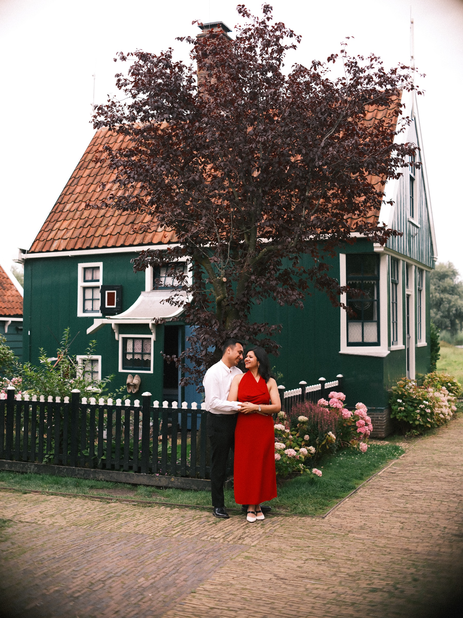 Green wooden house with an orange tile roof and a dark red-leaved tree, a couple standing small by the picket fence and hydrangeas