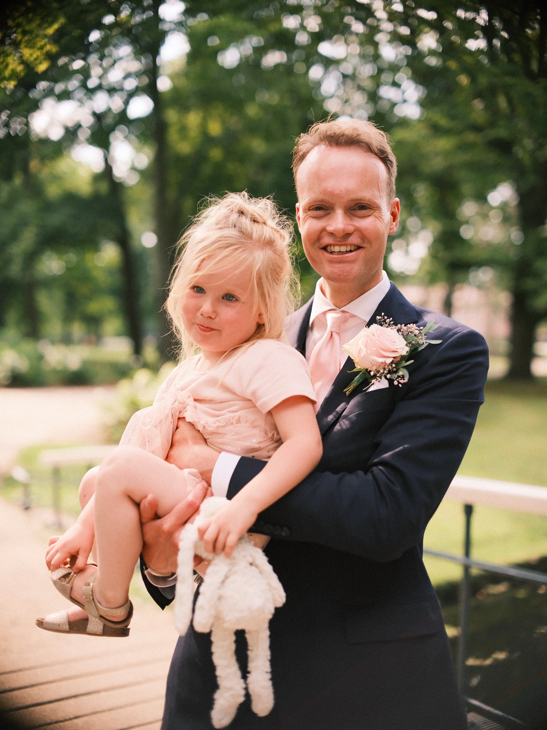 Bruidegom draagt een klein blond meisje met een zacht konijnenknuffeltje, allebei lachend op het bruggetje