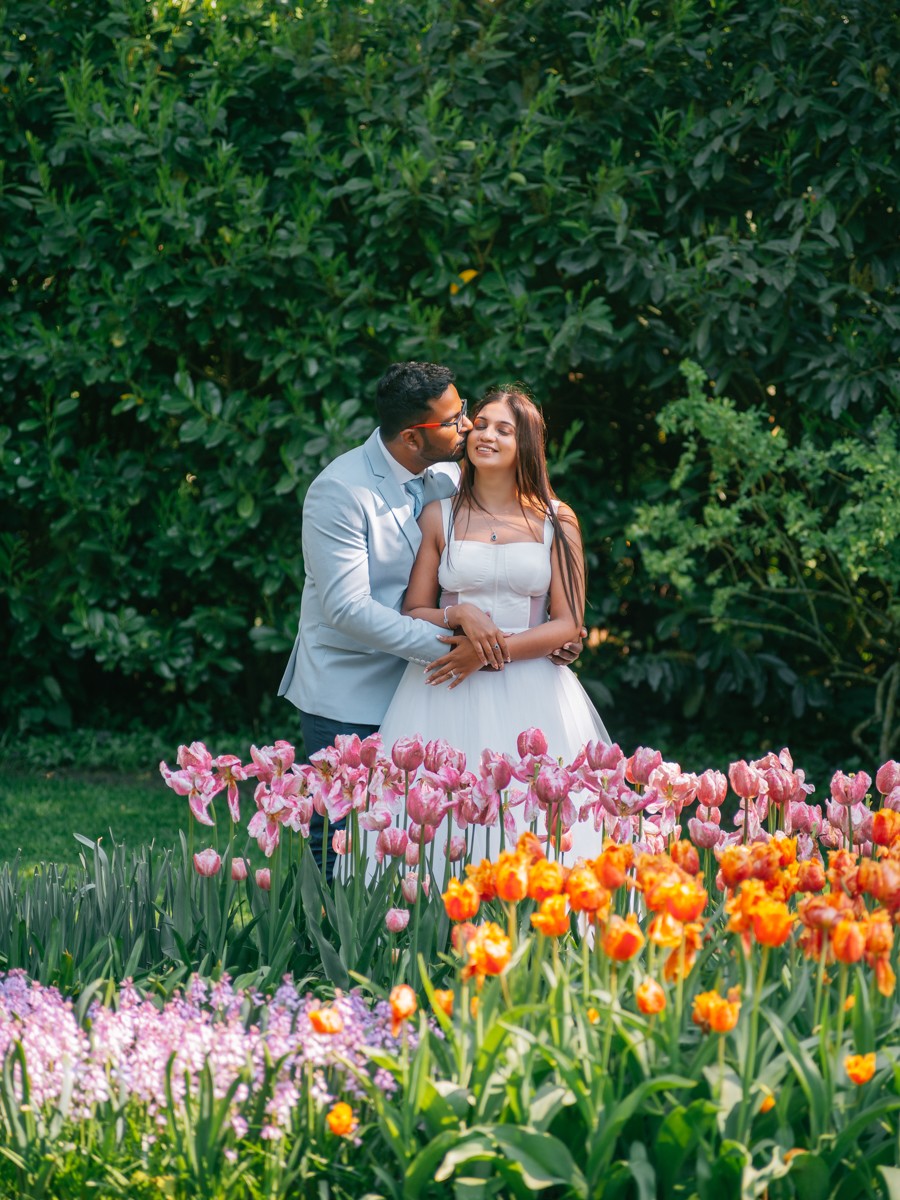 Bruidegom kust de bruid op de wang achter een perk met roze en oranje tulpen, met donkergroene struiken erachter