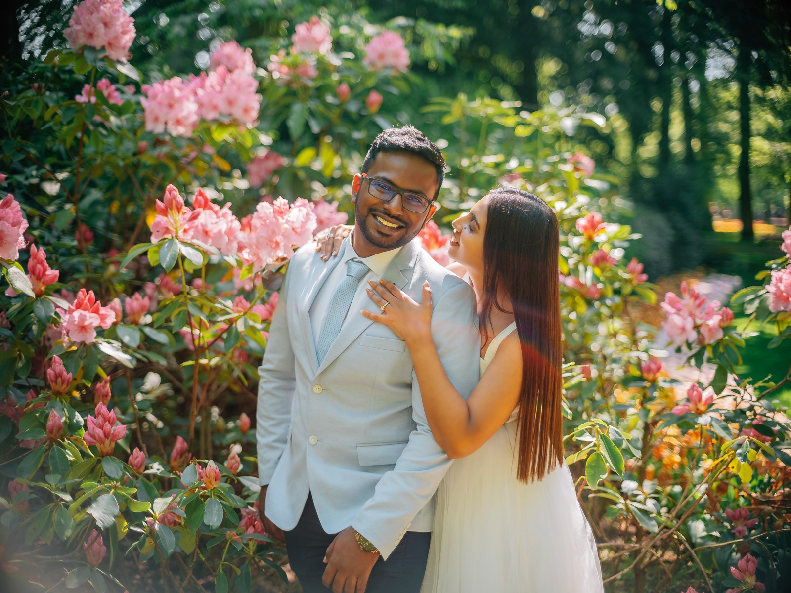 Bruidegom lacht voor roze rododendrons terwijl de bruid naar hem opkijkt, met één hand op zijn schouder