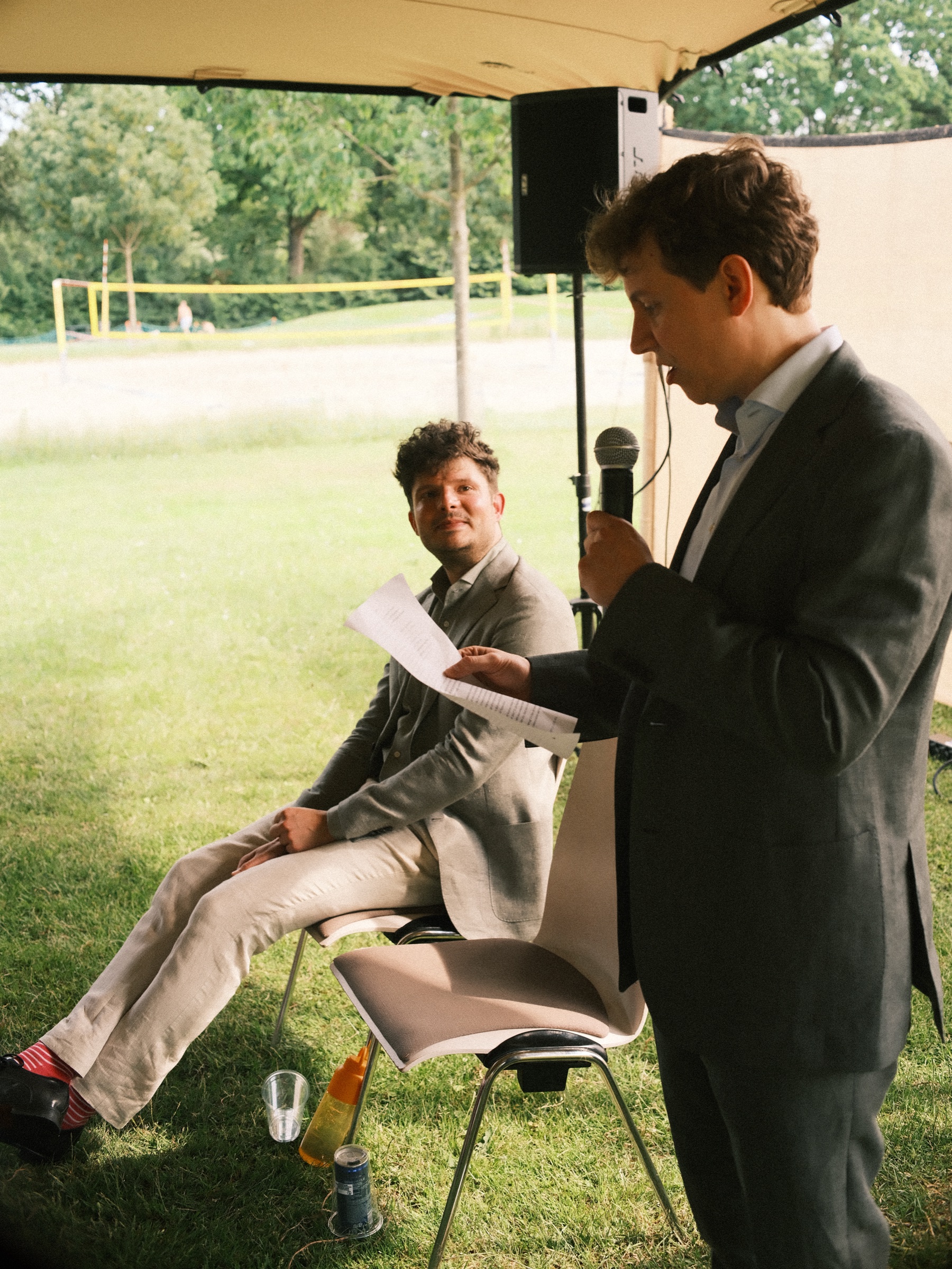 Groom in a gray suit reading his words into a microphone as the other groom, in a linen jacket and red socks, smiles up
