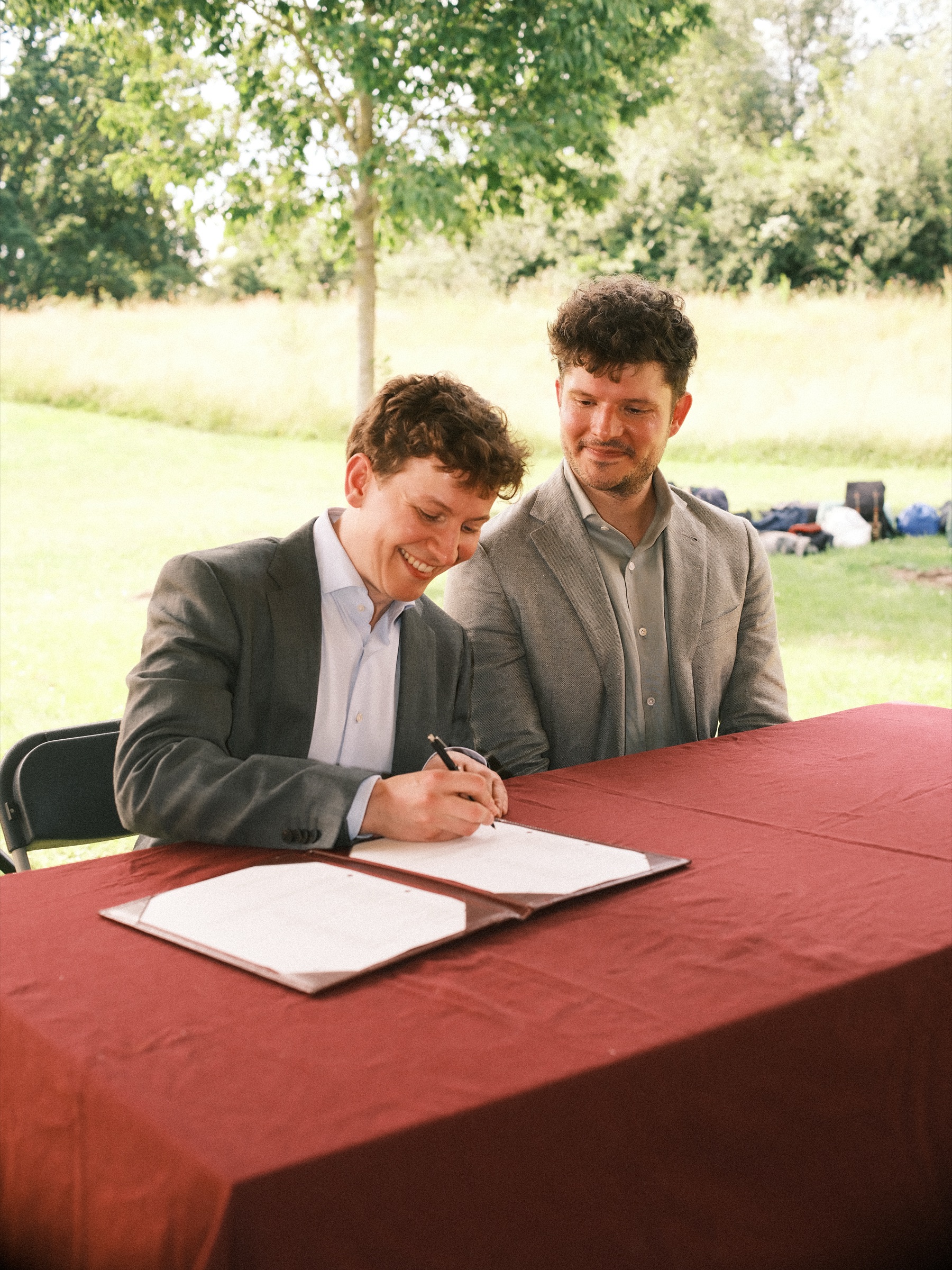 Smiling groom signing the marriage register at a table with a dark red cloth, his new husband laughing beside him