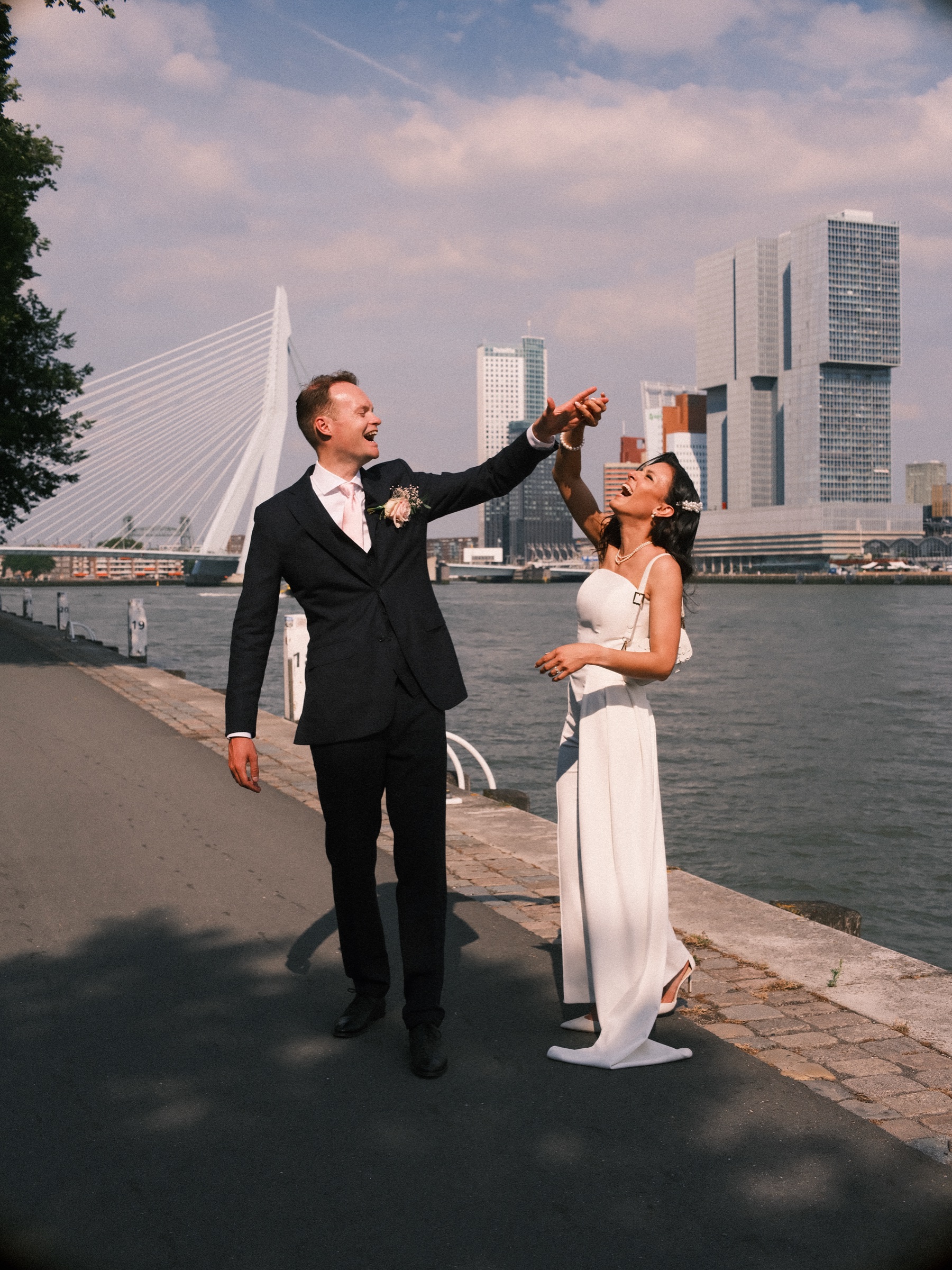 Bruidegom laat de lachende bruid in haar witte strapless jumpsuit draaien op de kade, met de Erasmusbrug erachter