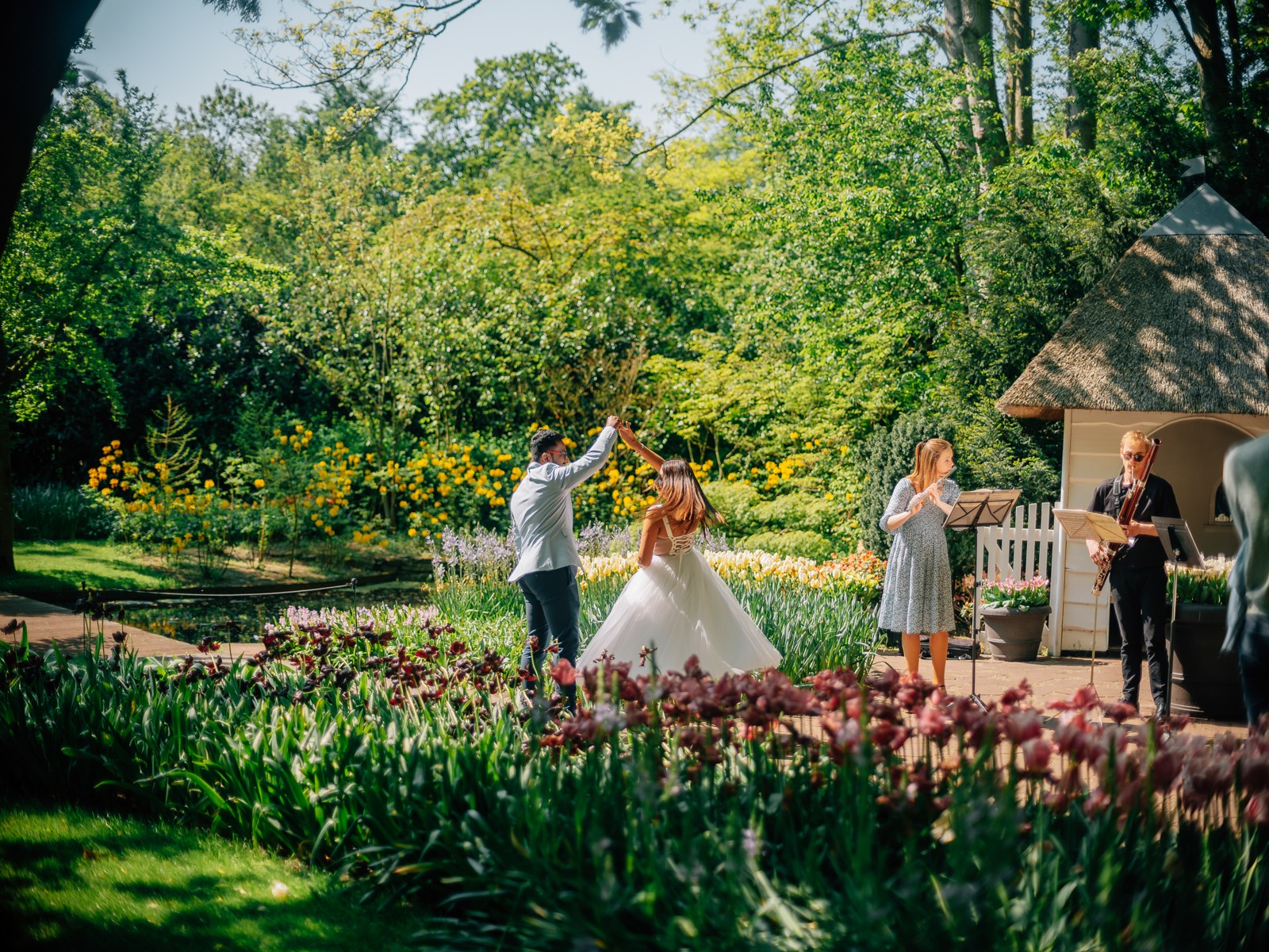 Bruidegom laat de bruid ronddraaien op een tuinpad terwijl een fluitist en een fagottist spelen bij een tuinhuisje in Keukenhof