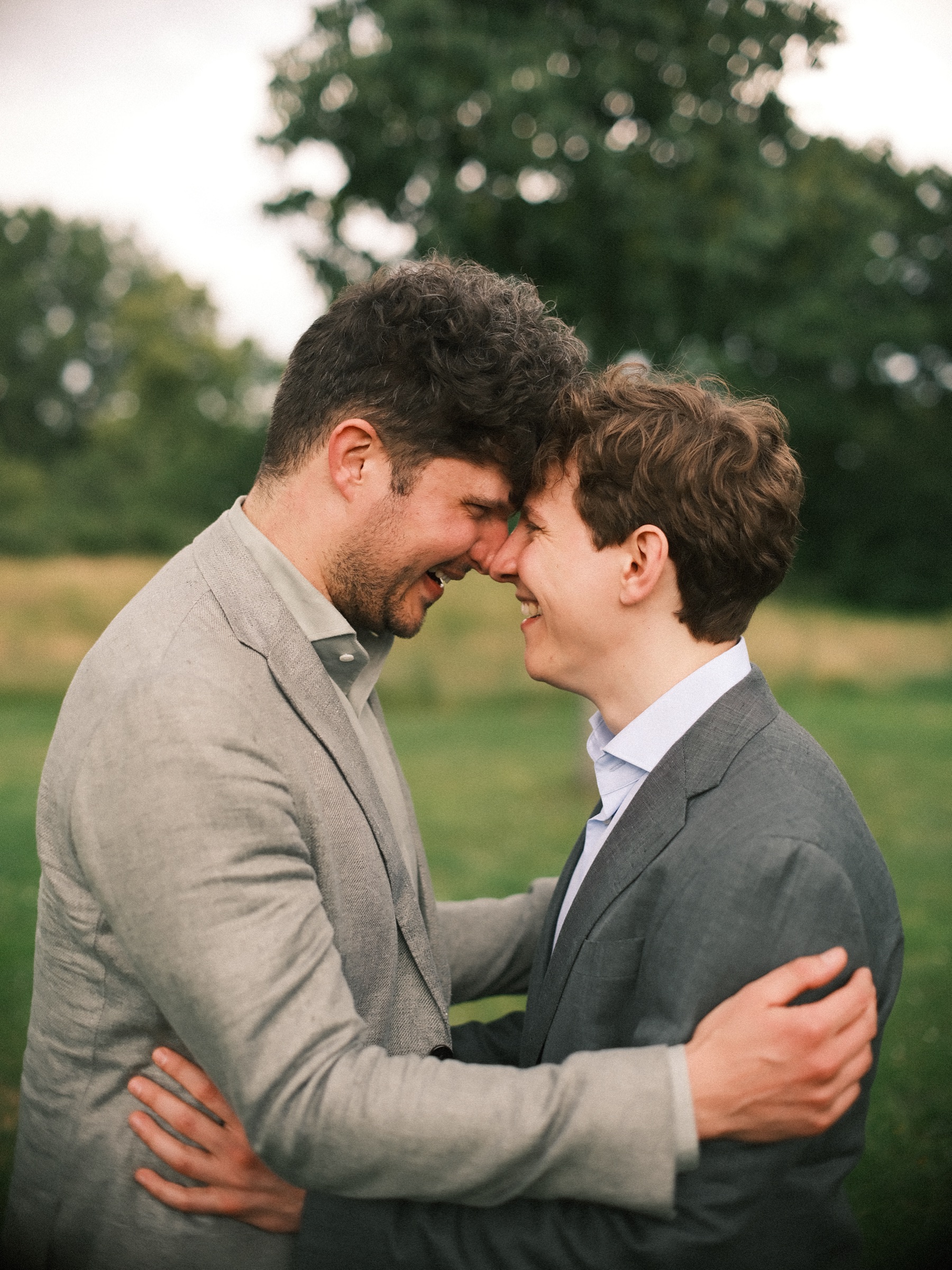 Newlywed grooms laughing with their foreheads together and their arms around each other on a green lawn in June