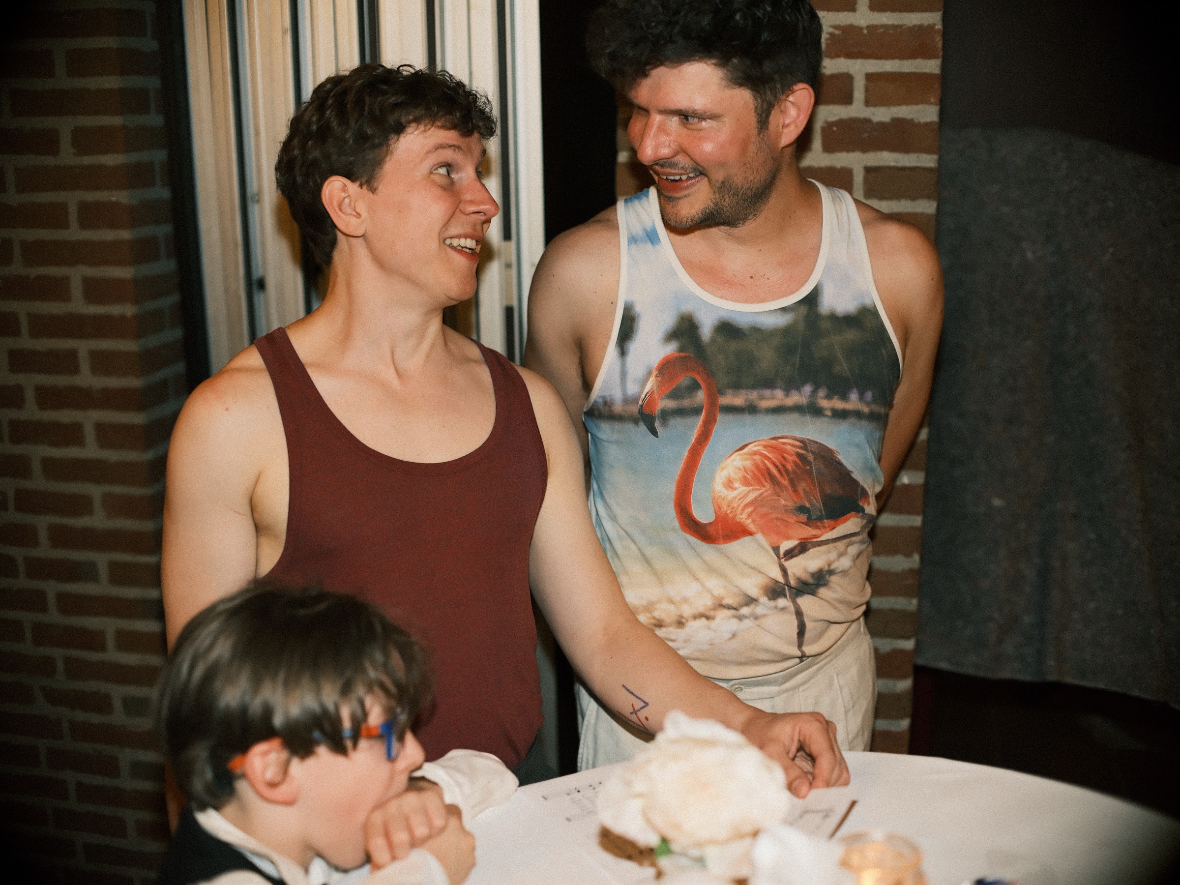Both grooms laughing in tank tops, one printed with a flamingo, at a table on the evening of their wedding in Utrecht