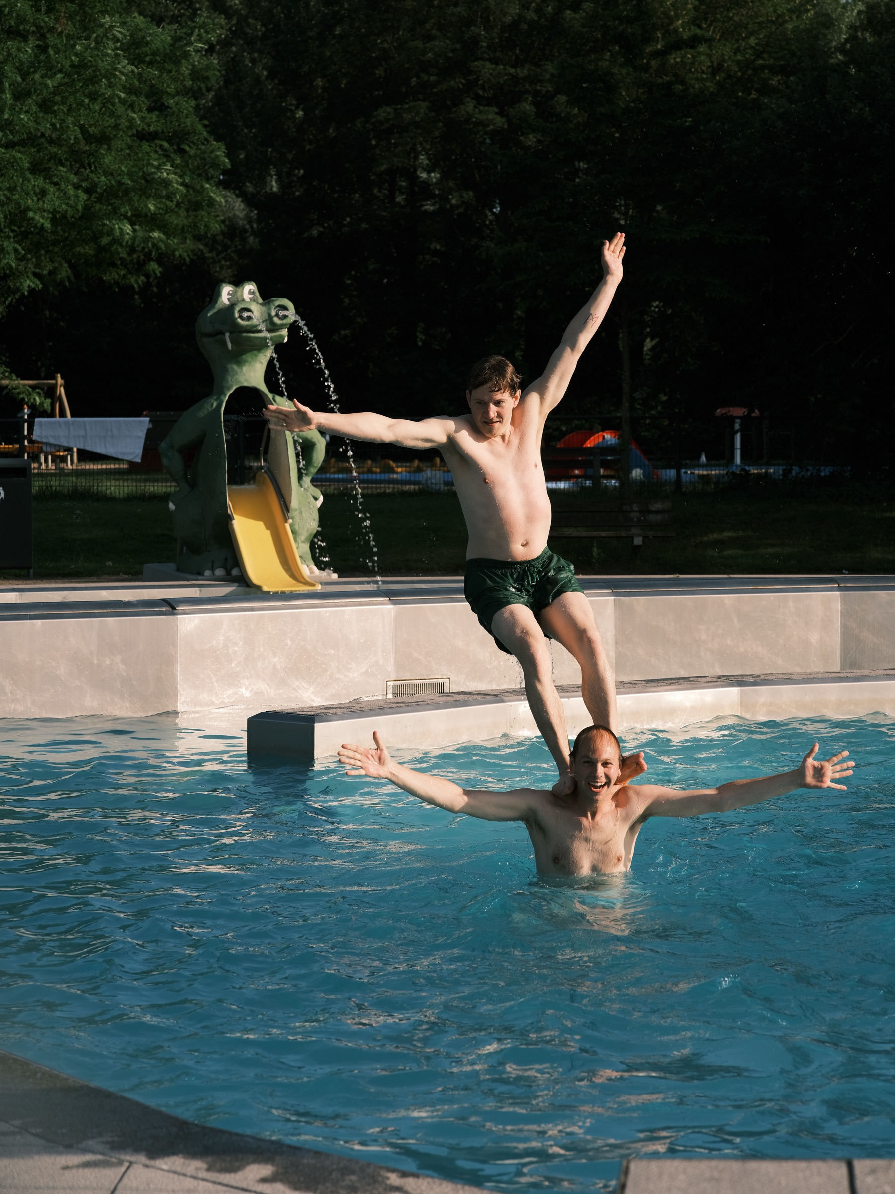 Guest jumping into an outdoor pool with arms spread past a green crocodile fountain, another guest cheering in the water