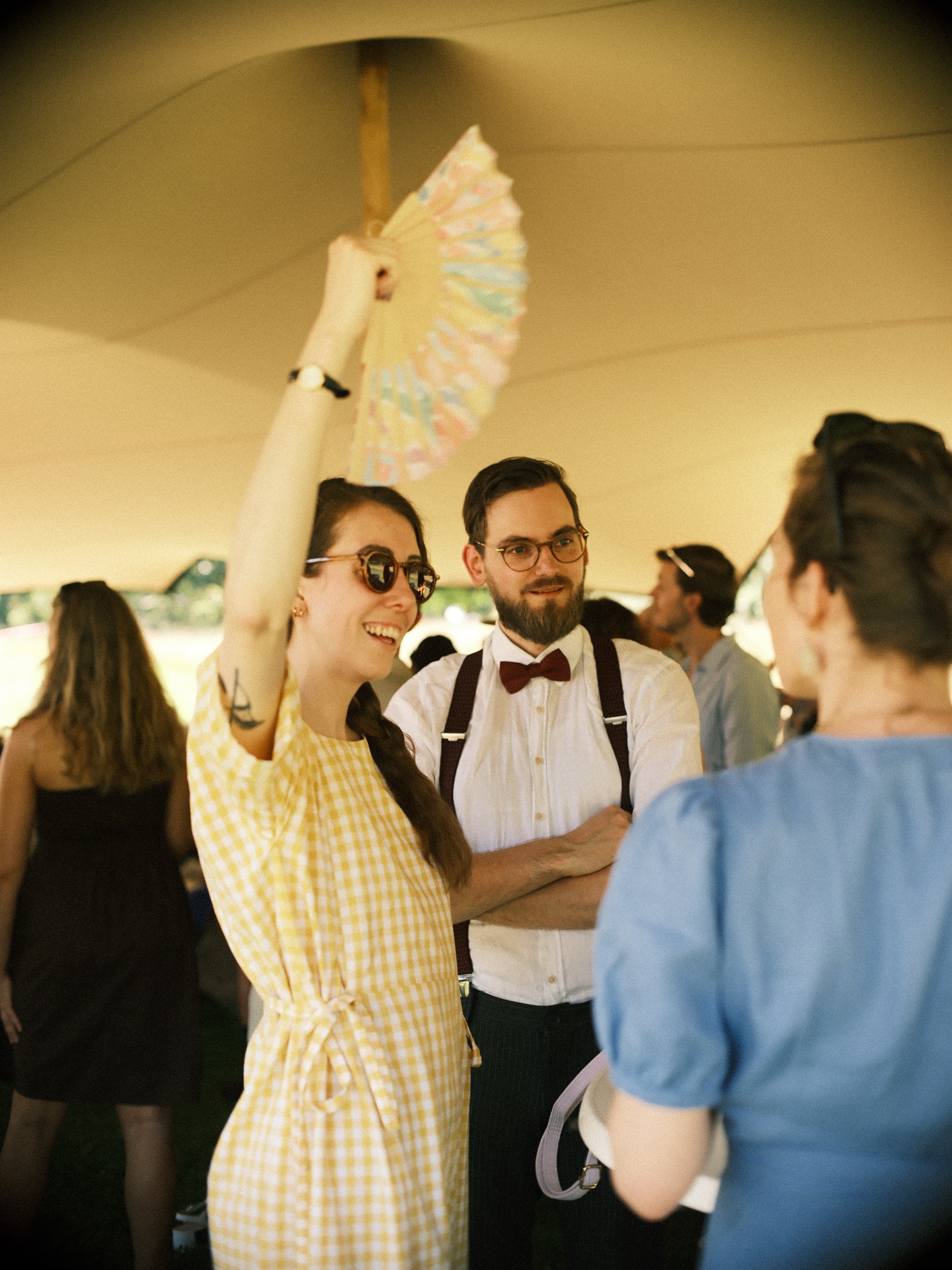 Laughing guest in a yellow gingham dress and sunglasses waving a pastel paper fan, a man in a red bow tie beside her