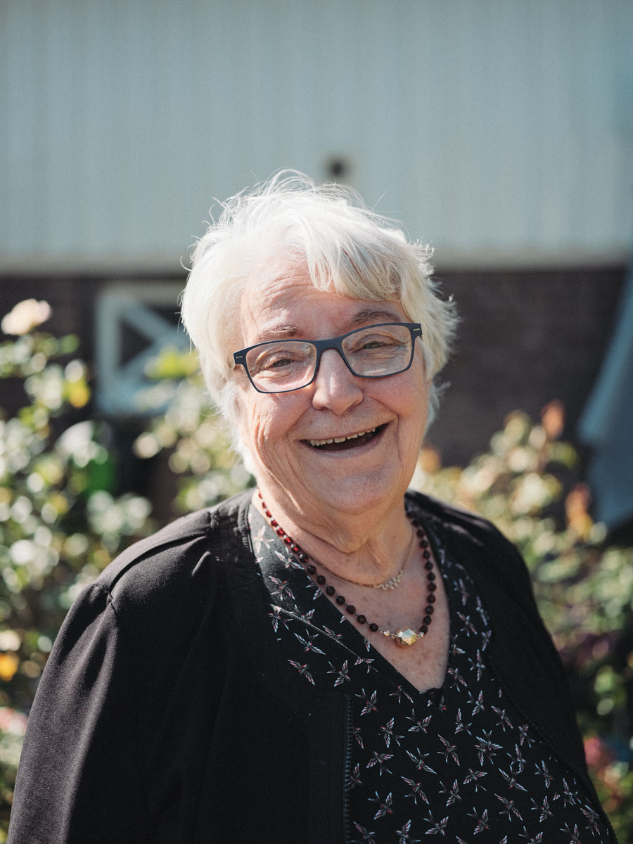 Older guest with white hair and glasses laughing in the sunshine, a dark cardigan and a string of red beads around her neck