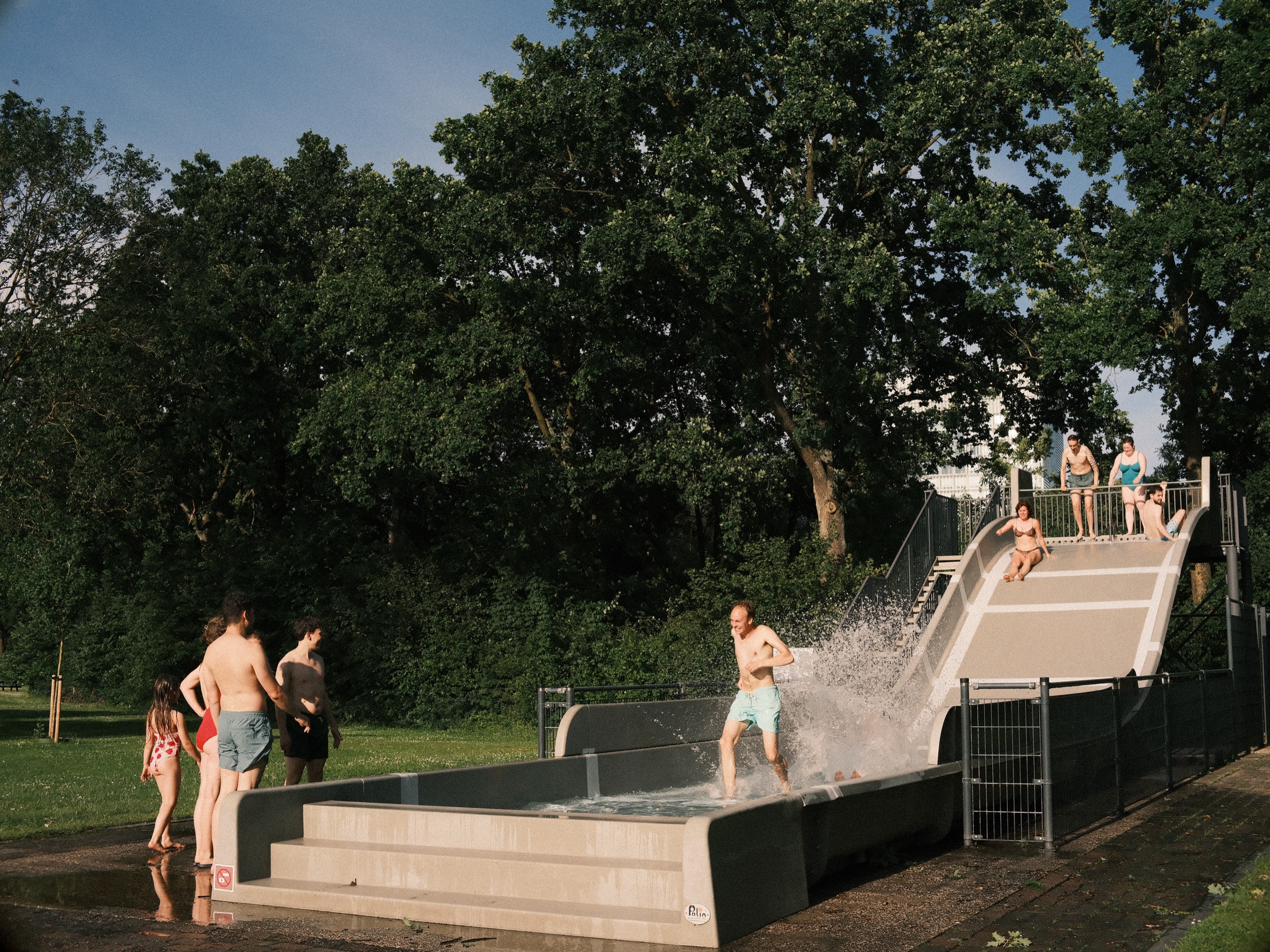 Guest splashing down at the foot of a wide water slide in front of tall dark green trees, others in swimwear watching