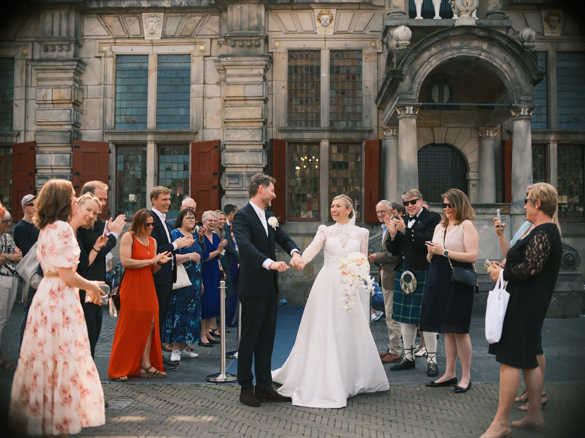 Bruidspaar hand in hand op de Markt in Delft terwijl de gasten juichen, onder wie een man in een kilt en een vrouw in het oranje