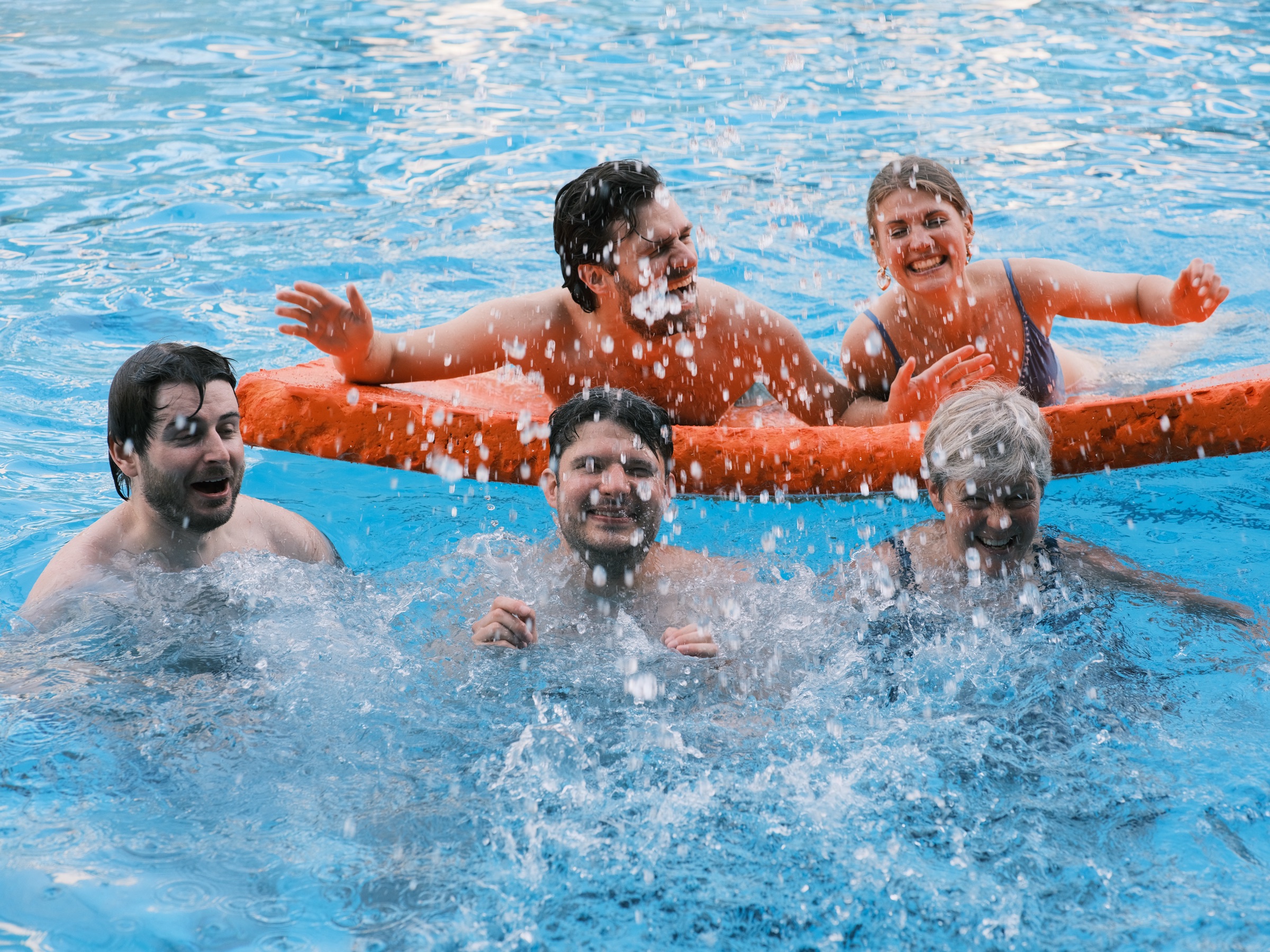 Four guests and an older woman splashing around a bright orange float in the blue pool, water drops flying through the air