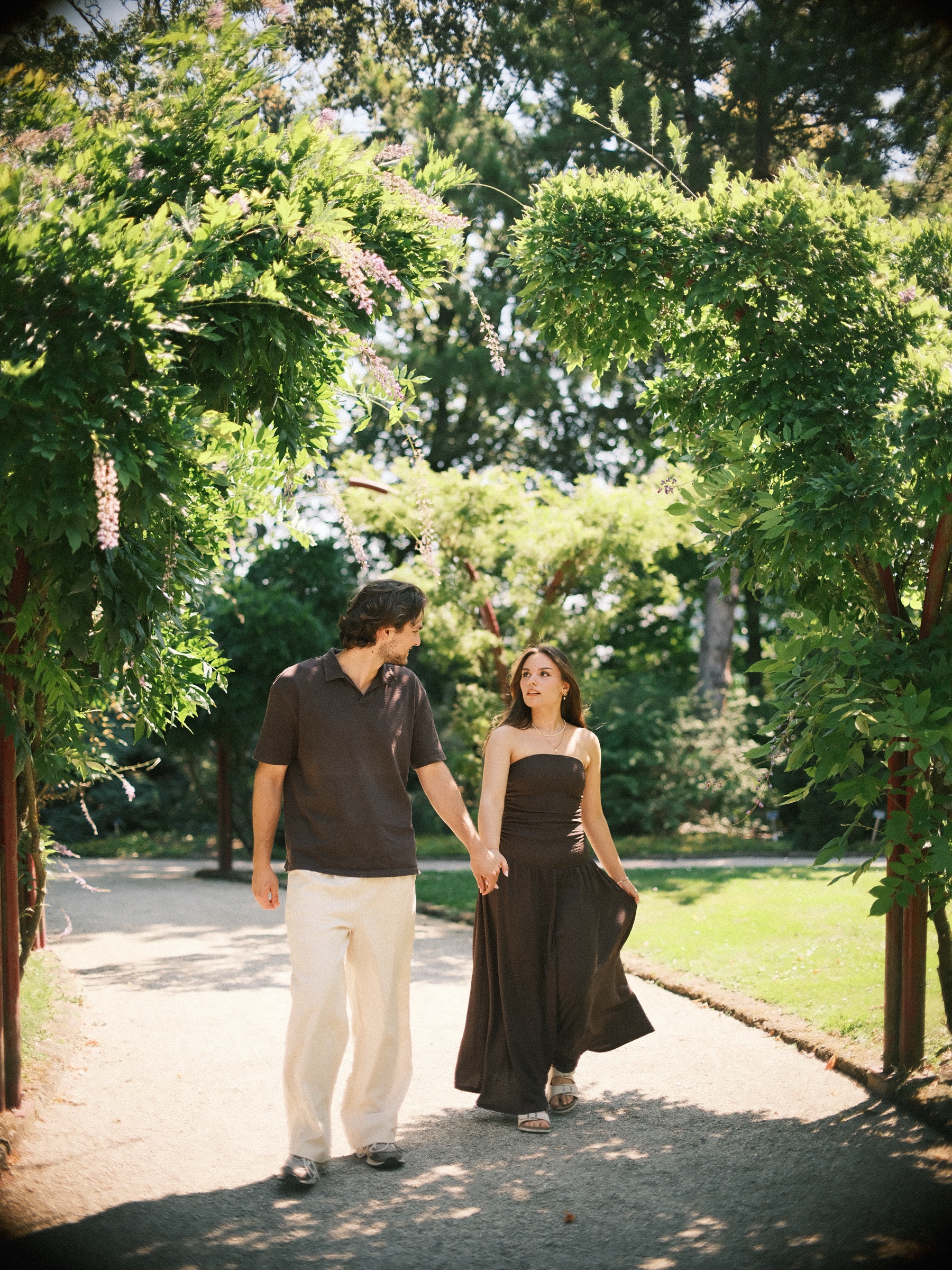Walking hand in hand under a green arch on a gravel path, he in cream trousers and she in a long brown dress, in the July sun