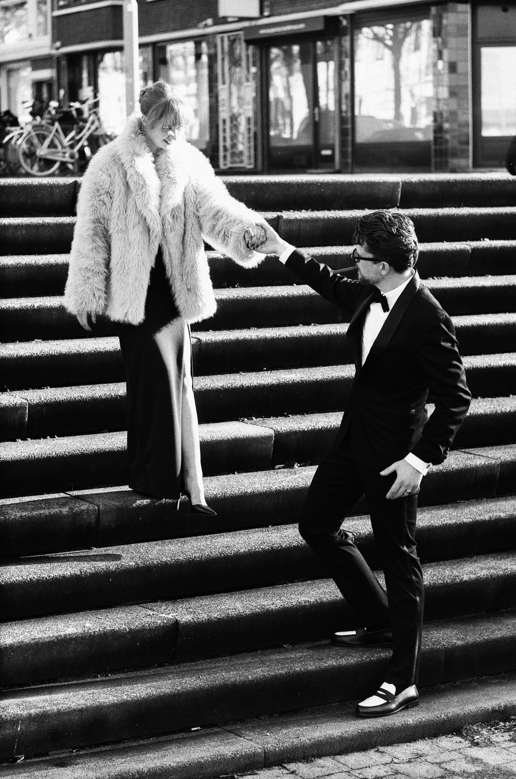 On wide stone steps in Rotterdam he bends to kiss her hand as she walks down in a long slit dress and fur coat, long shadows on film