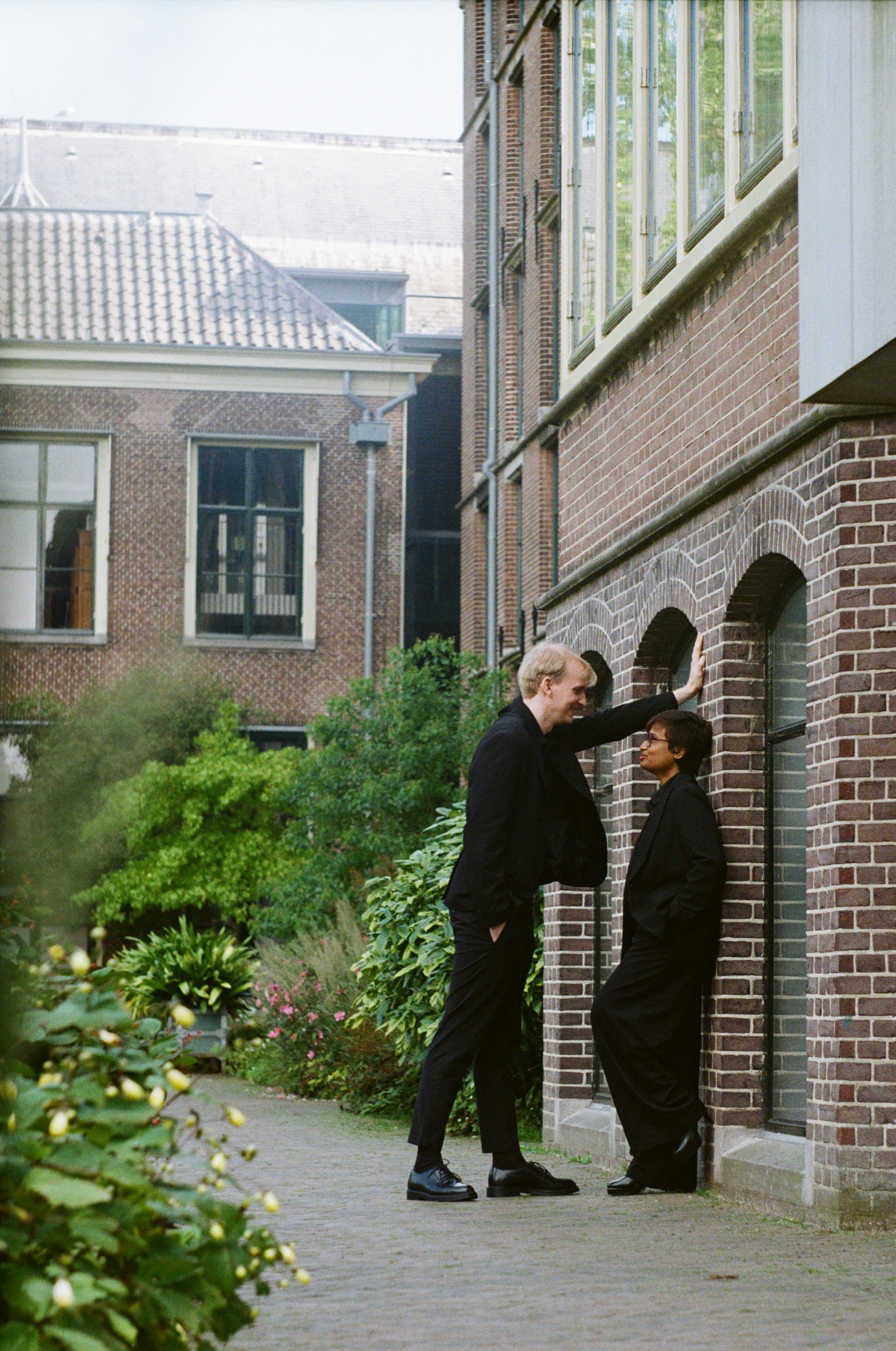 He leans one hand on a brick wall above her under a row of arches, green garden plants and yellow flowers along the path