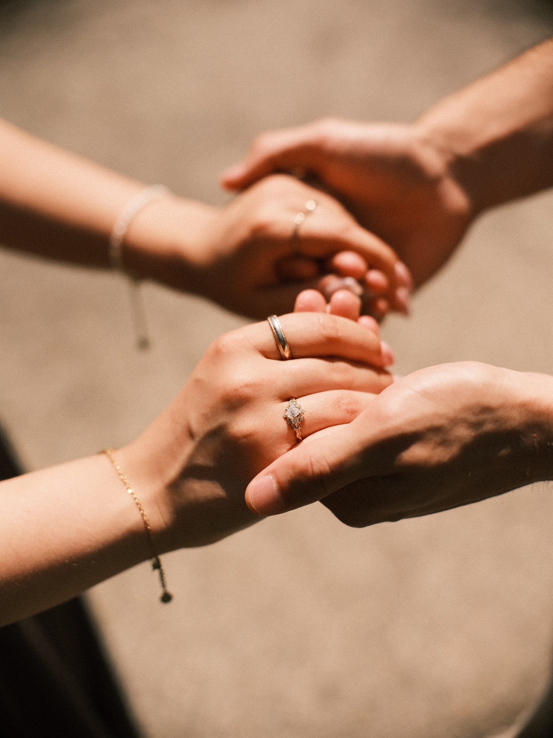 Close view of hands held together in warm light, her engagement ring and thin bracelets in the middle, the gravel path a soft blur