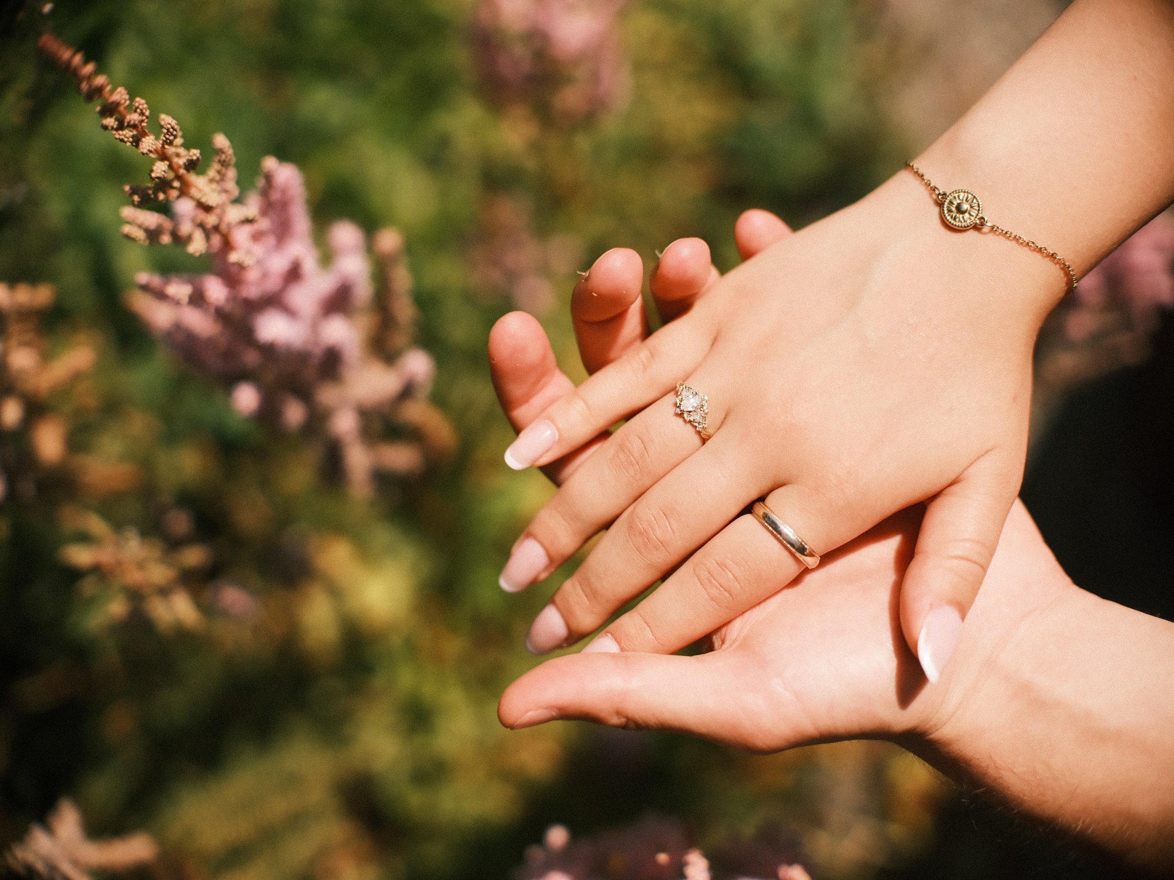 Her hand with the engagement ring and a gold bracelet resting on his palm, a feathery pink plume beside them in the garden
