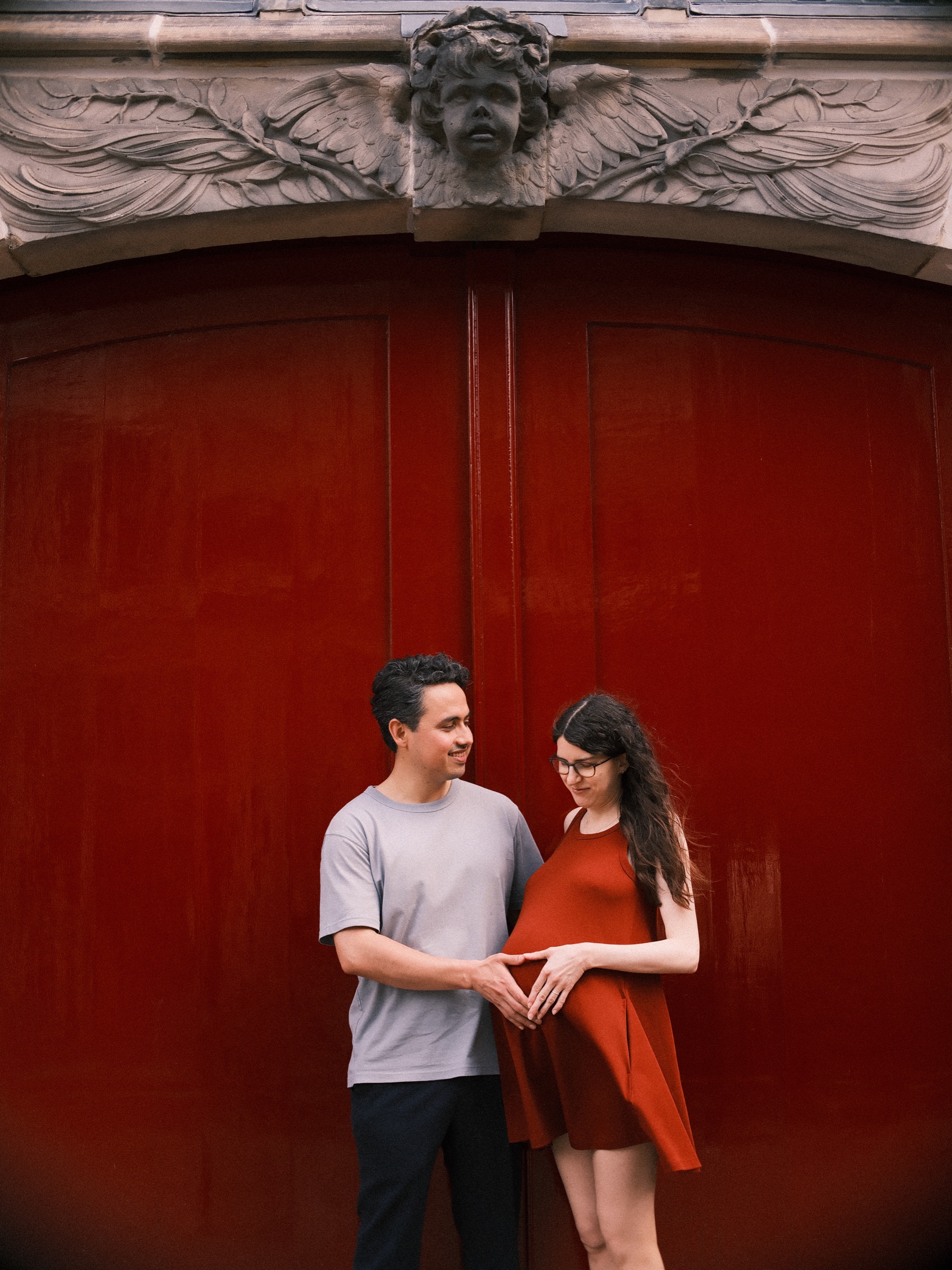 Couple shaping a heart with their hands around her baby bump in front of a deep red door, his grey T-shirt beside her dress