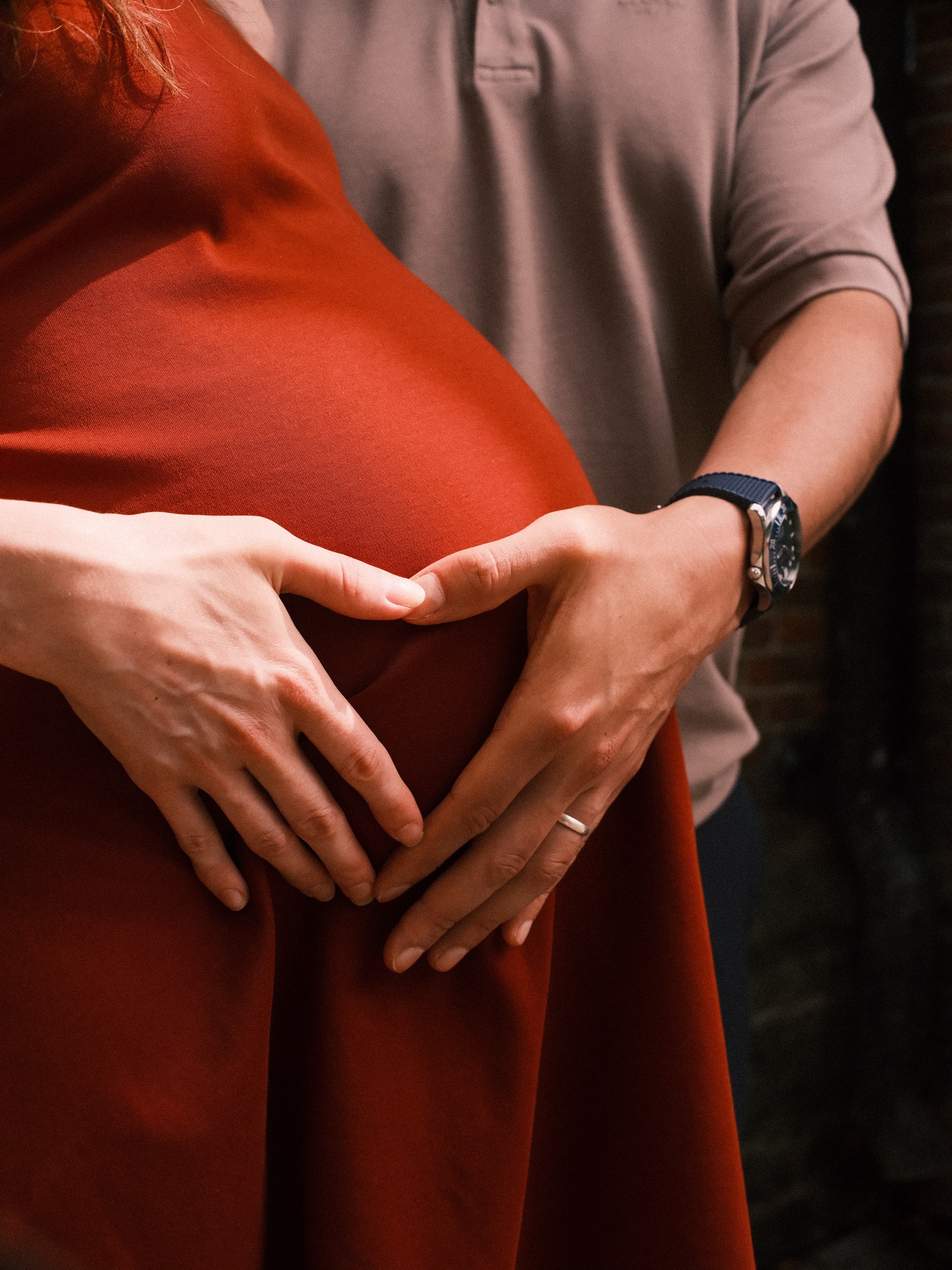 Two hands forming a heart on a baby bump in a rust-red dress, a ring on her finger and a watch on his wrist