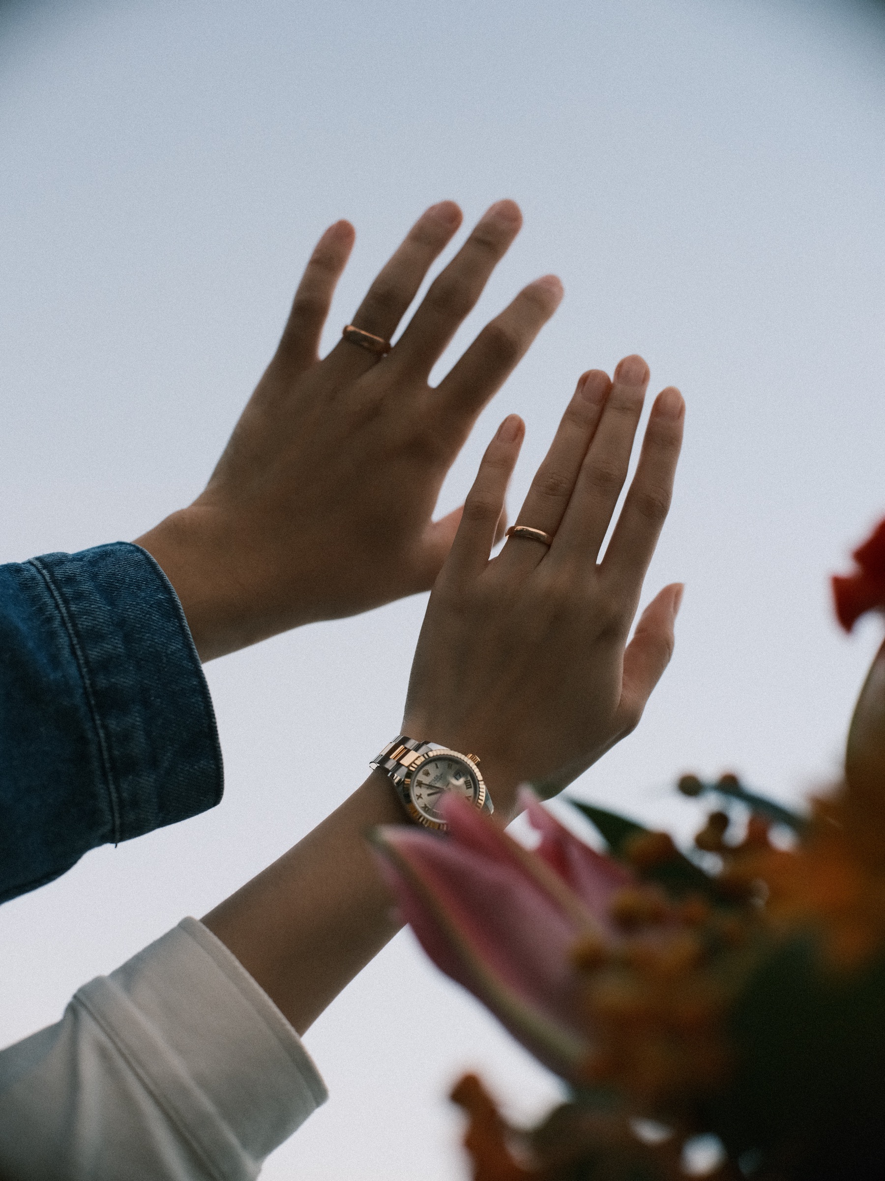 Twee opgeheven handen met dezelfde gouden ringen tegen een bleke lucht, een tweekleurig horloge om haar pols