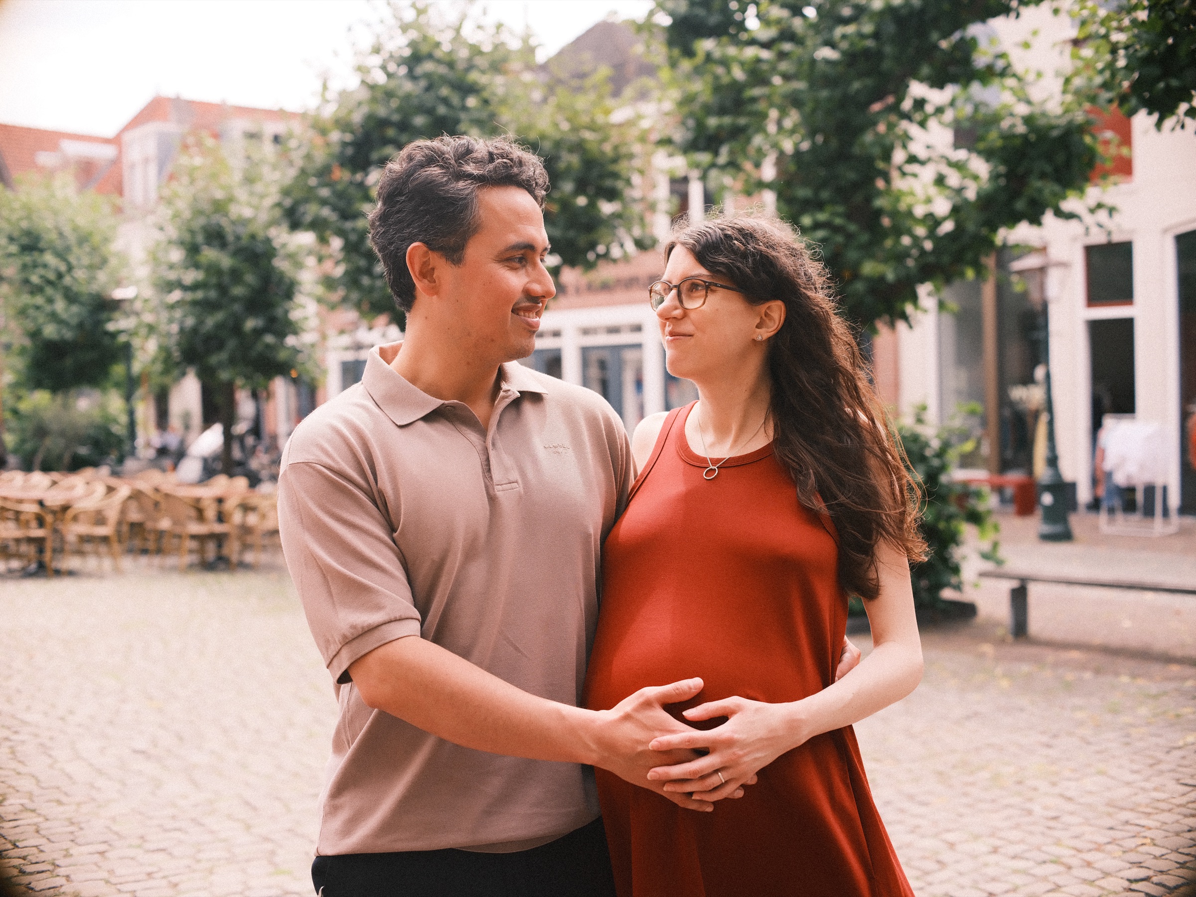He looks at her and smiles as their hands meet over her baby bump, café terraces and green trees on a sunny square in Leiden