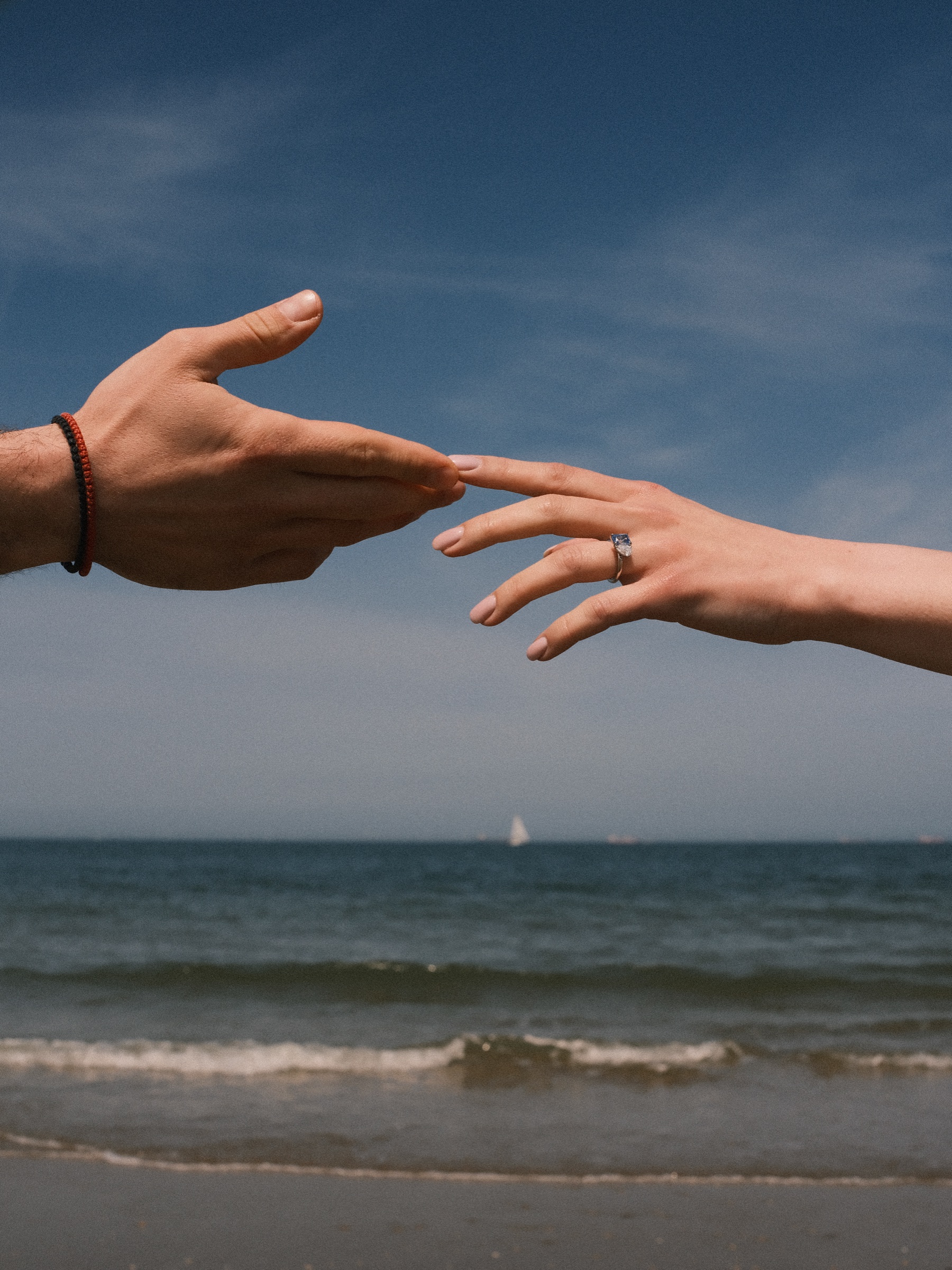 Two hands reaching for each other above the sea, her new ring catching the light and a small sailboat on the horizon