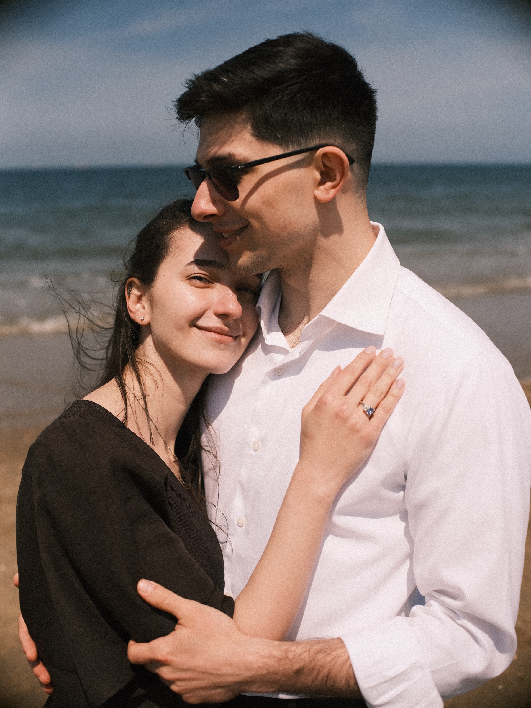 She leans her head against his chest and smiles, her ring hand on his white shirt, while he looks down at her by the sea