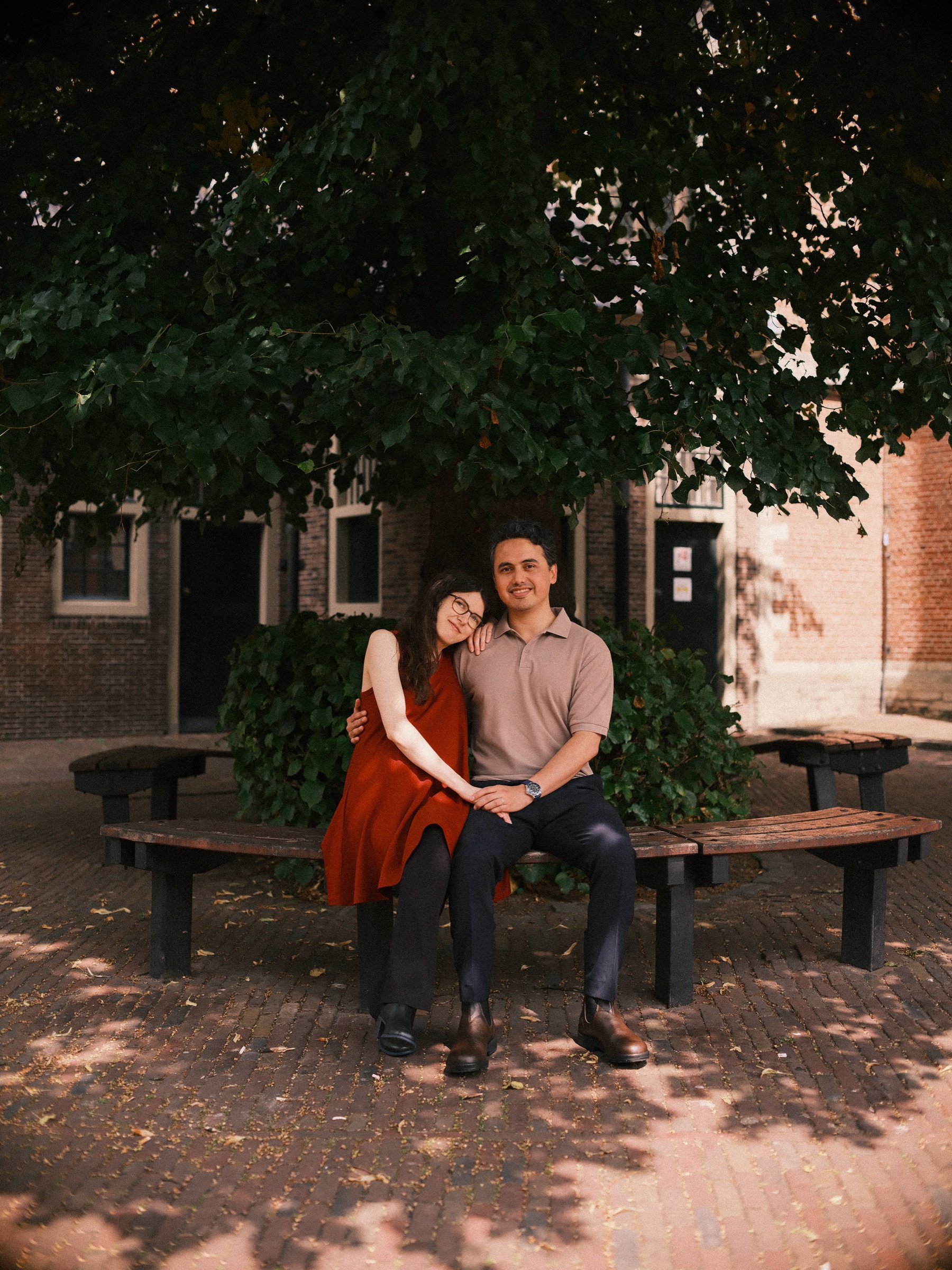 Pregnant woman in a rust-red dress resting her head on her partner's shoulder on a round wooden bench in deep shade
